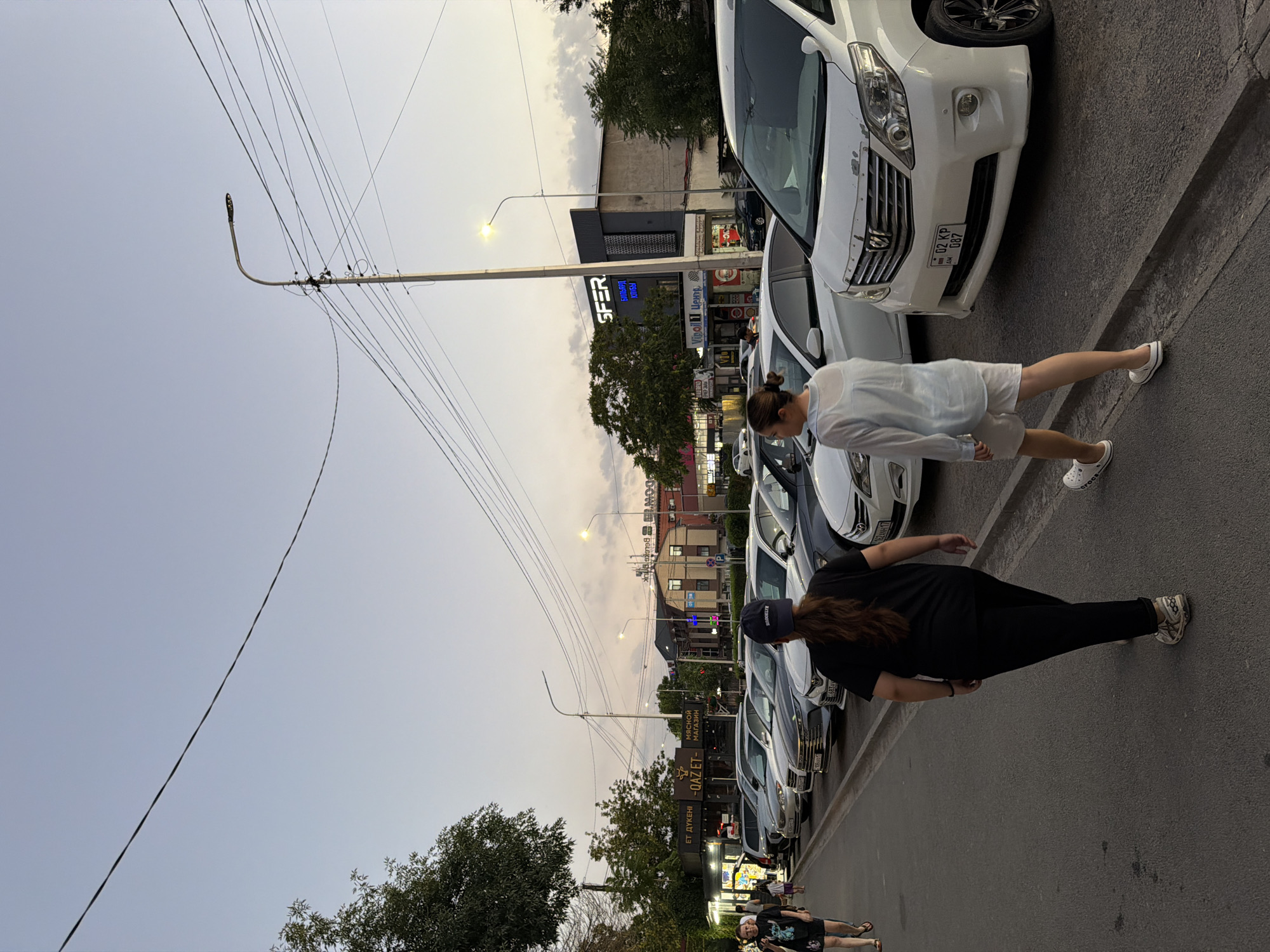 The image depicts a street scene in Shymkent, Kazakhstan, likely during dusk given the soft, diffused light and visible streetlights. The perspective is from ground level, looking up and across the street, with the image rotated 90 degrees counter-clockwise.
Two women are the primary human subjects. One, wearing a light blue shirt and white shorts, is walking away from the viewer and toward the parked cars. The second woman, dressed in a black t-shirt and black leggings, is also walking away from the viewer, slightly ahead of the first woman. She is wearing a dark baseball cap.
Several white and silver cars are parked along the side of the street. One white car in the foreground has a license plate visible that reads "02 KP 087" and appears to be from Kazakhstan. A building with illuminated signs stands behind the parked cars. Some of the visible text on the signs includes "SFER", "БИЛЬЯРД", "КЛУБЫ", "ЕТ ДҮКЕНІ", "МЯСНОЙ МАГАЗИН", and "Vipoj 1 Центр".
Utility poles with numerous overhead wires are prominent in the scene, stretching across the sky, which is a pale, overcast grey with hints of yellow or orange near the horizon. Trees with green foliage are also visible, adding to the urban but naturalistic setting. The overall atmosphere appears calm and ordinary, suggesting a typical evening on a city street.
The image is rich with details of urban life, including transportation, commercial establishments, and infrastructure, set against a soft twilight sky.