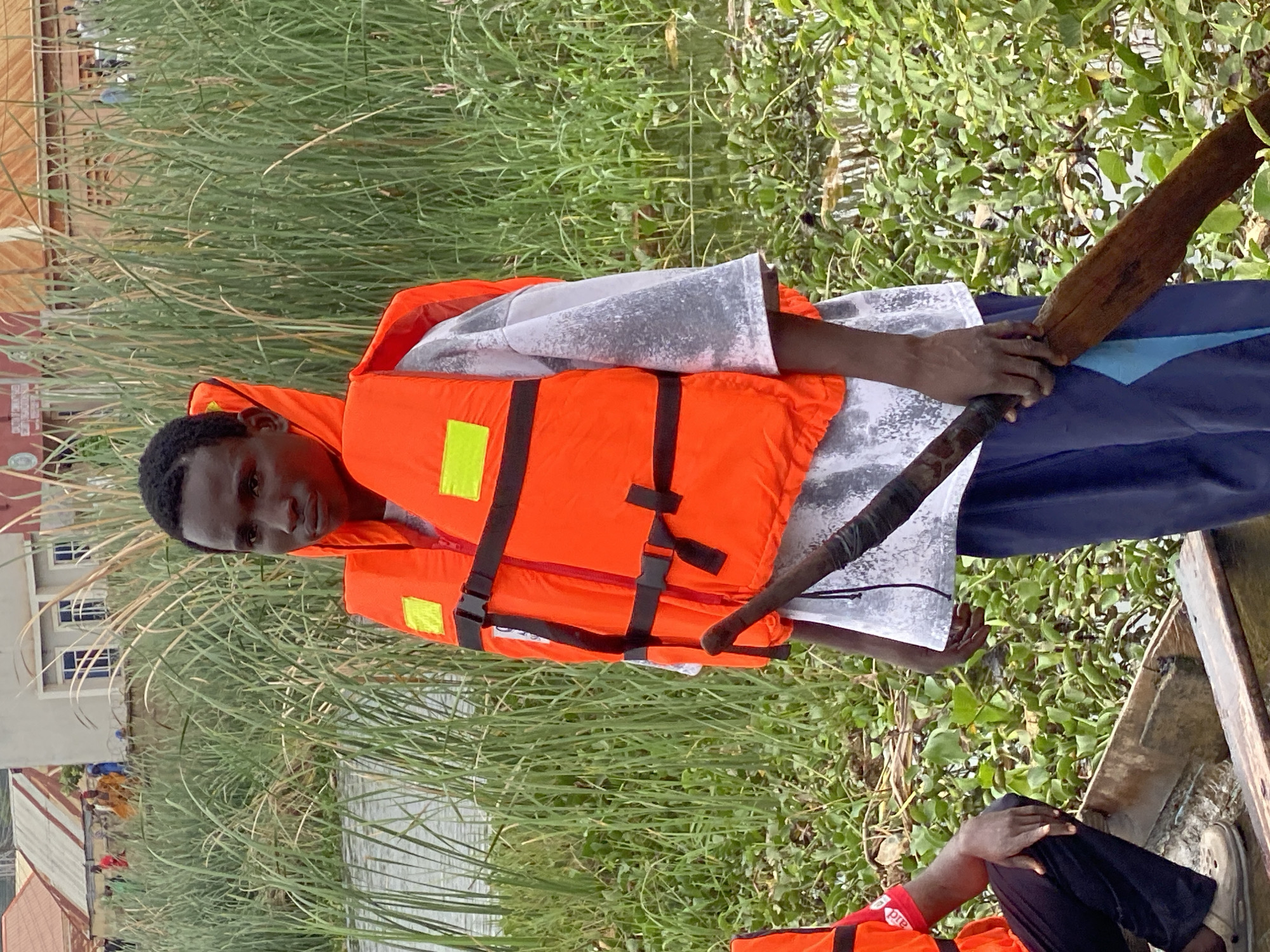 The image shows a young Black male wearing an orange life vest over a grey t-shirt and blue pants. He is standing in a wooden boat, holding a wooden oar. Behind him, there is a body of water with dense green vegetation and reeds. In the background, there are buildings with red and white roofs, suggesting an urban or semi-urban setting. People can be seen in the background near the buildings. The overall environment appears to be a waterfront area in Kawo, Nigeria.