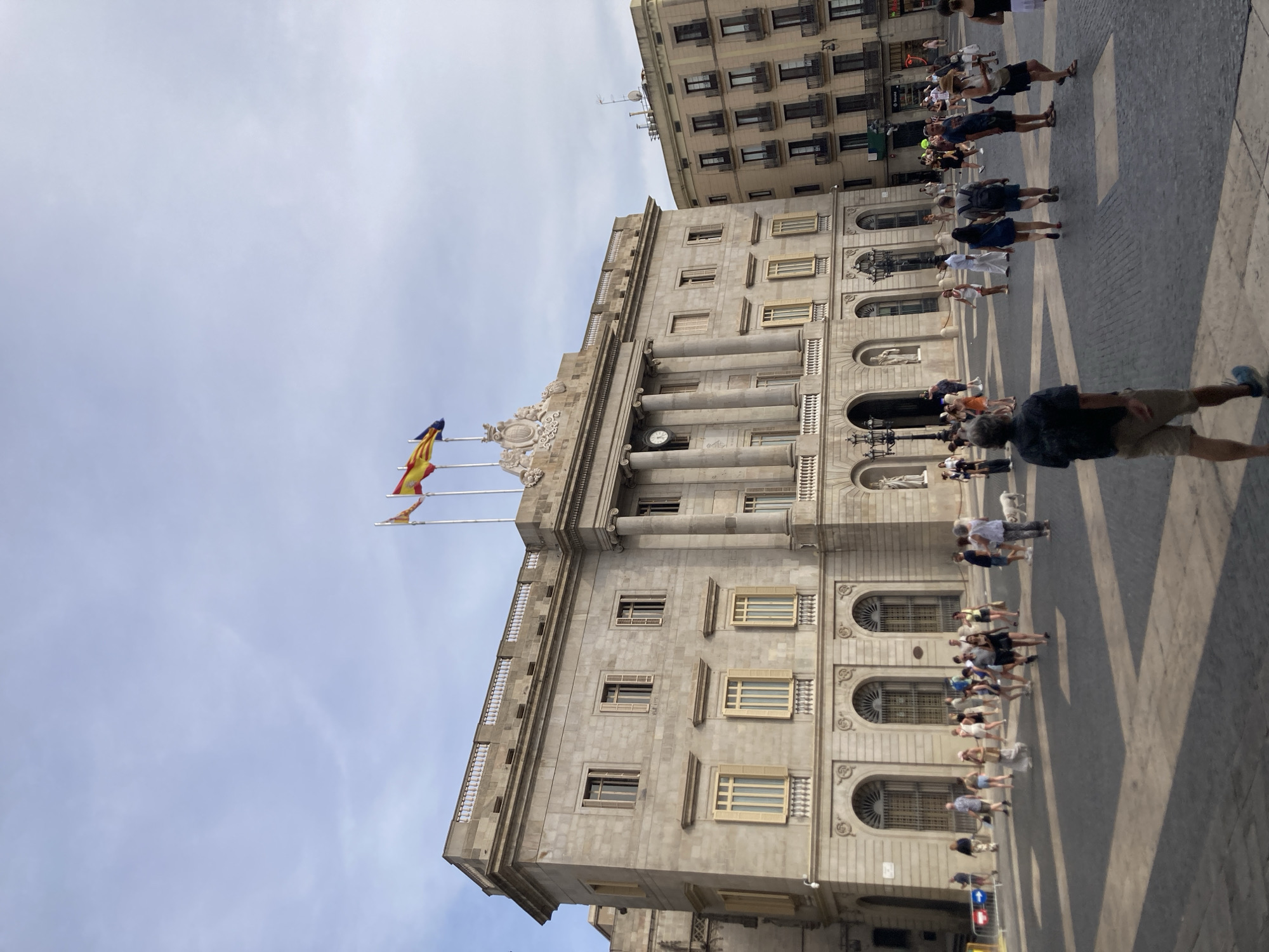 The image shows a view of the Barcelona City Hall (Ajuntament de Barcelona) on a clear day. The building's facade is made of stone with classical architectural elements, including columns, arches, and decorative moldings. Several flags, including the Spanish flag, are visible flying from poles on the building.
In the foreground and midground, a paved plaza is filled with many people. They are casually dressed, suggesting a warm day, and appear to be walking, standing, and interacting with each other. The shadows cast by the people and buildings indicate it is likely late morning or early afternoon.
The overall scene is bustling and vibrant, capturing a typical day in a public square in a European city. The architecture suggests a historic and significant location. The presence of numerous people suggests this is a popular tourist destination or a central gathering place. No discernible text is visible in the image.