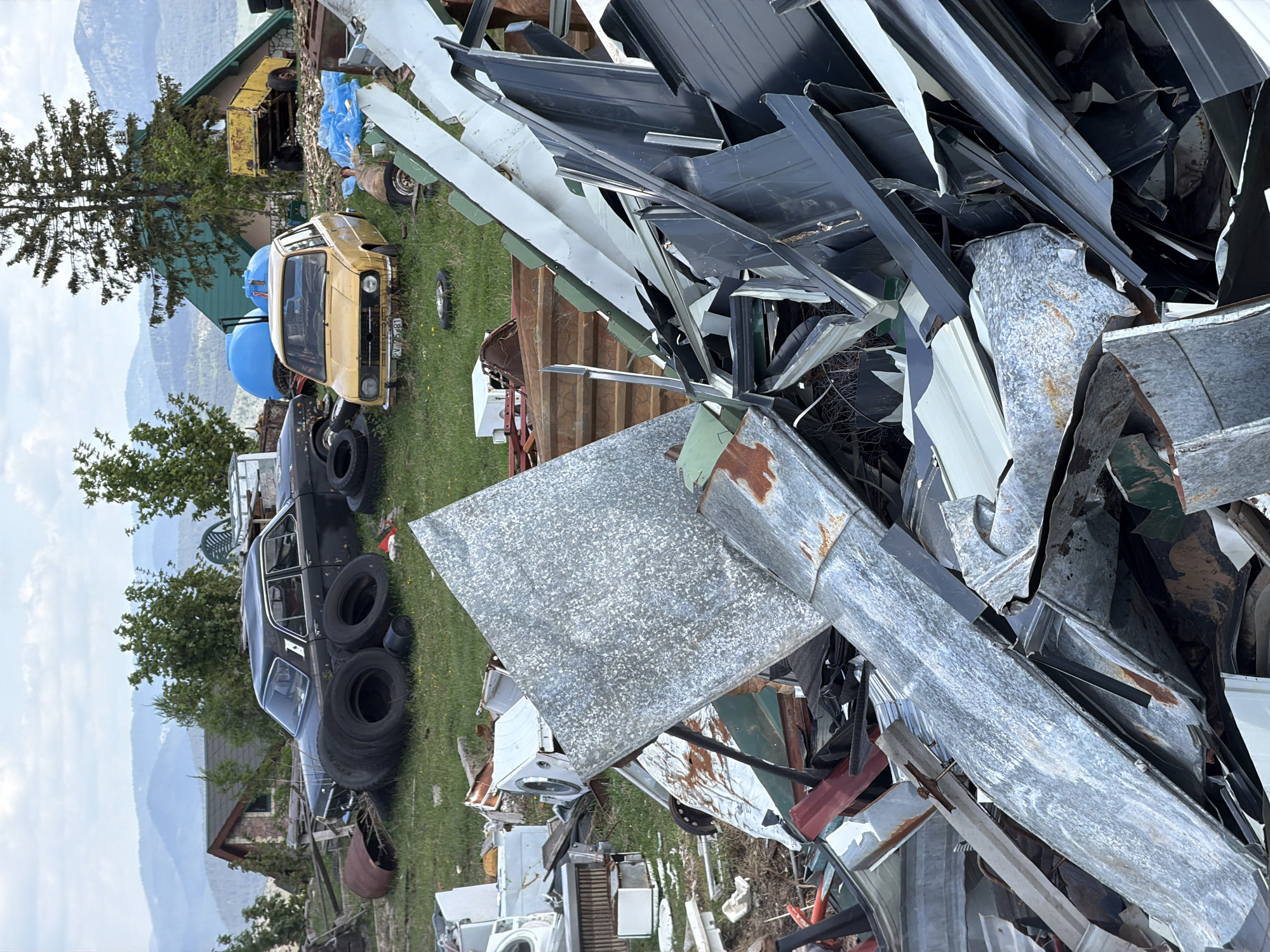 The image shows a cluttered scene, likely in a rural area of Žabljak, Montenegro. A jumble of scrap metal, including sheets of galvanized metal, black metal pieces, and other unidentified metal objects, dominates the foreground. In the background, partially obscured, are abandoned or derelict vehicles, including a yellow car and a black car with tires stacked on its roof. Other discarded items such as appliances (washing machines) are also visible. The overall impression is one of neglect and uncontrolled waste accumulation. The landscape includes grassy areas, trees, and distant mountains under an overcast sky.