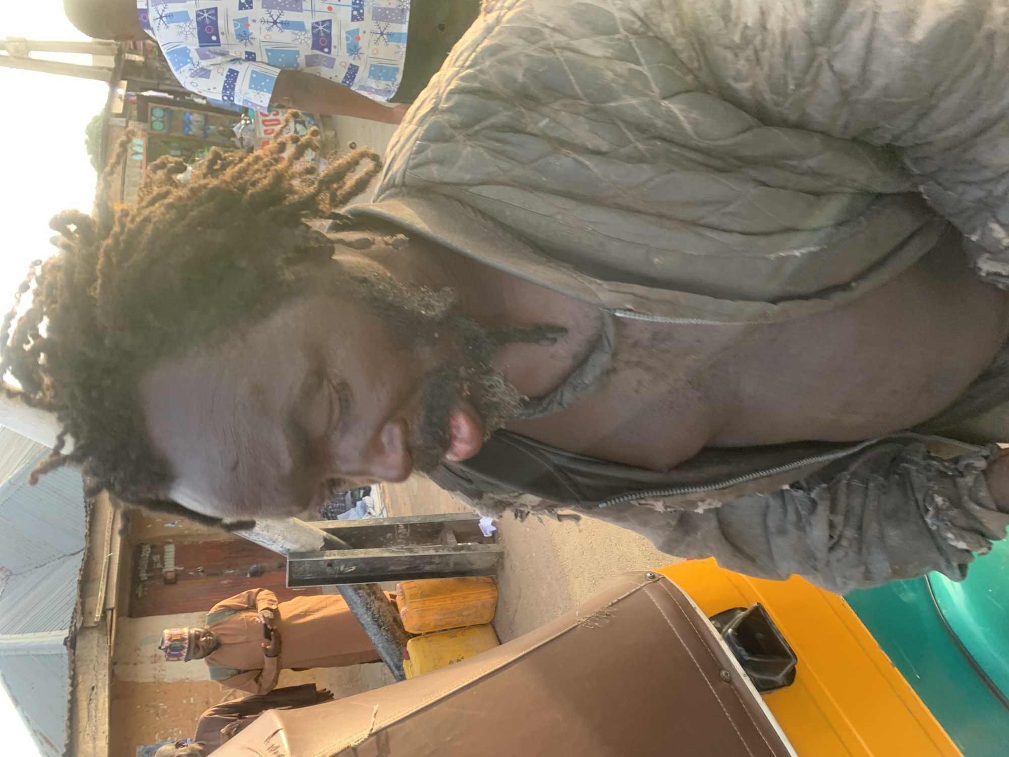 A man with dreadlocks and a beard wearing a torn, unzipped jacket stands partially inside a three-wheeled vehicle. The vehicle has a brown roof, a yellow body, and a green side. In the background, an older man wearing a brown robe and a patterned hat sits on a bench. Several yellow containers are visible near the bench. Another man wearing a shirt with a white and blue pattern is partially visible in the top left corner of the frame. The scene appears to be in a city street. This image may have been taken in Jos, Nigeria.