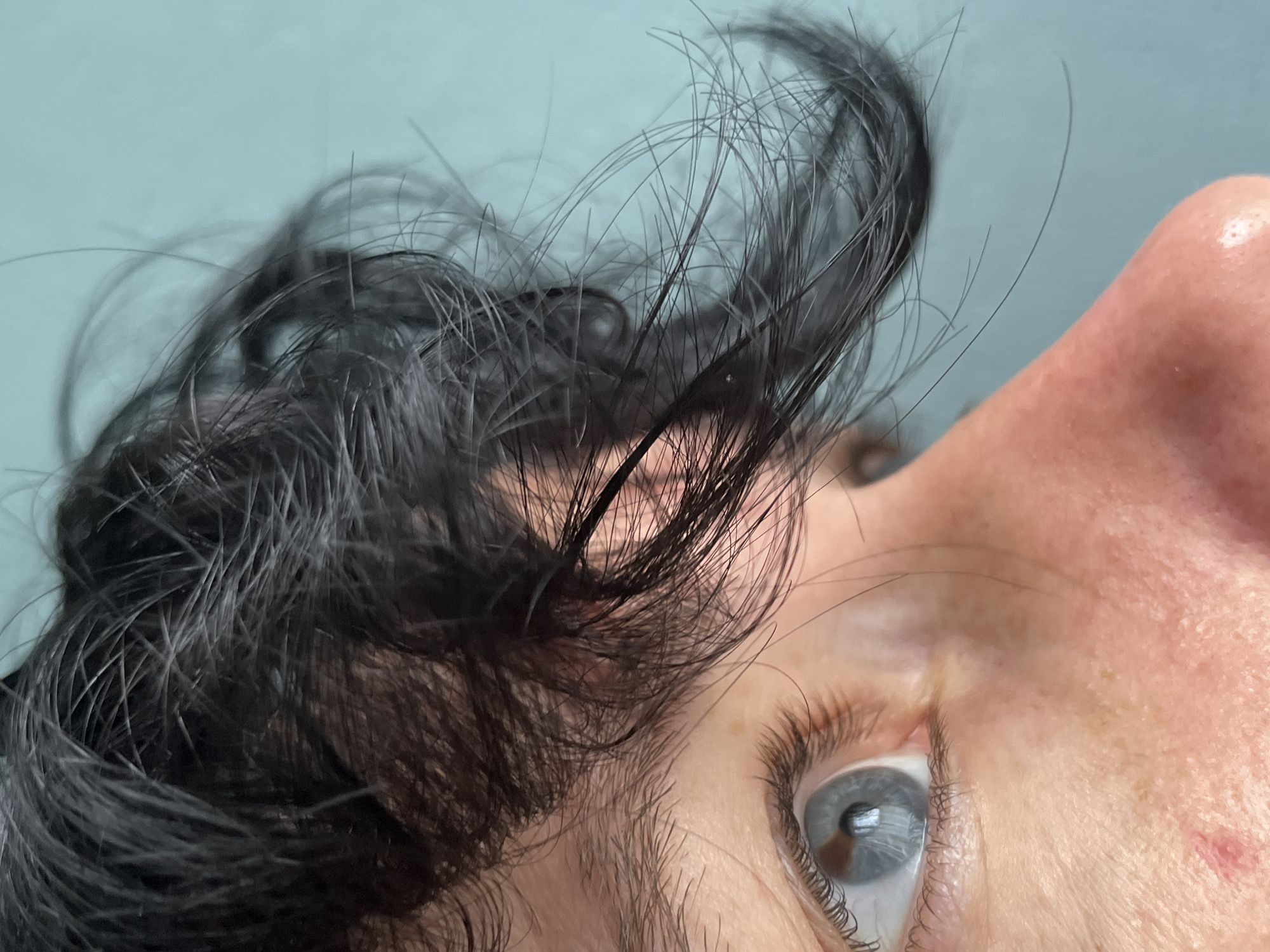 The close-up, eye-level shot shows the left side of a person's face, including their eye, nose, and dark, messy hair. The person has light skin with some pinkish undertones and a small reddish mark below the eye. Their eye is light blue with visible eyelashes and reflections in the pupil. The background is a solid, light teal color. The image does not depict a specific location in the city of ღრმაღელის თემი, Georgia, as it is a close-up portrait.