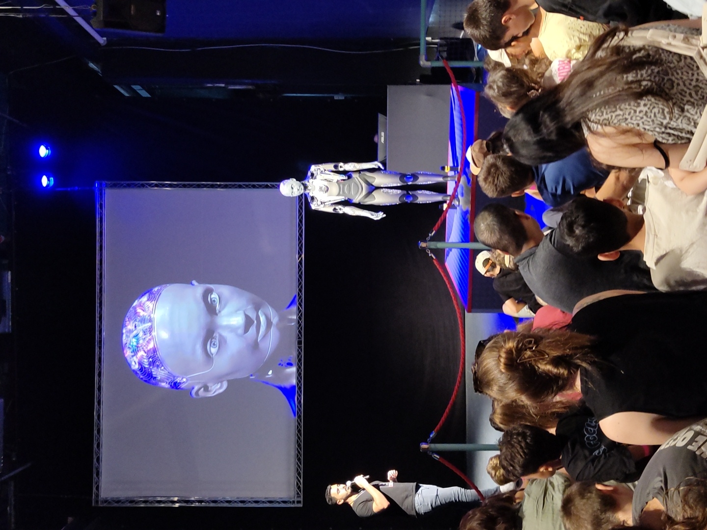 The image depicts an indoor event, likely a presentation or demonstration, where a humanoid robot is on display. In the foreground, a crowd of people, mostly facing away from the camera, are seated and watching the presentation. In the mid-ground, a man in a black t-shirt and jeans is speaking into a microphone, standing to the left of a large screen. The screen displays a close-up image of a robot's head. To the right of the screen, a standing humanoid robot is visible, wearing a silver and white suit. The background consists of a dark stage with blue lighting. The presence of red rope barriers suggests a controlled viewing area. While the image itself does not provide explicit geographical markers for Haifa, Israel, the event is described as taking place in Haifa, Israel.