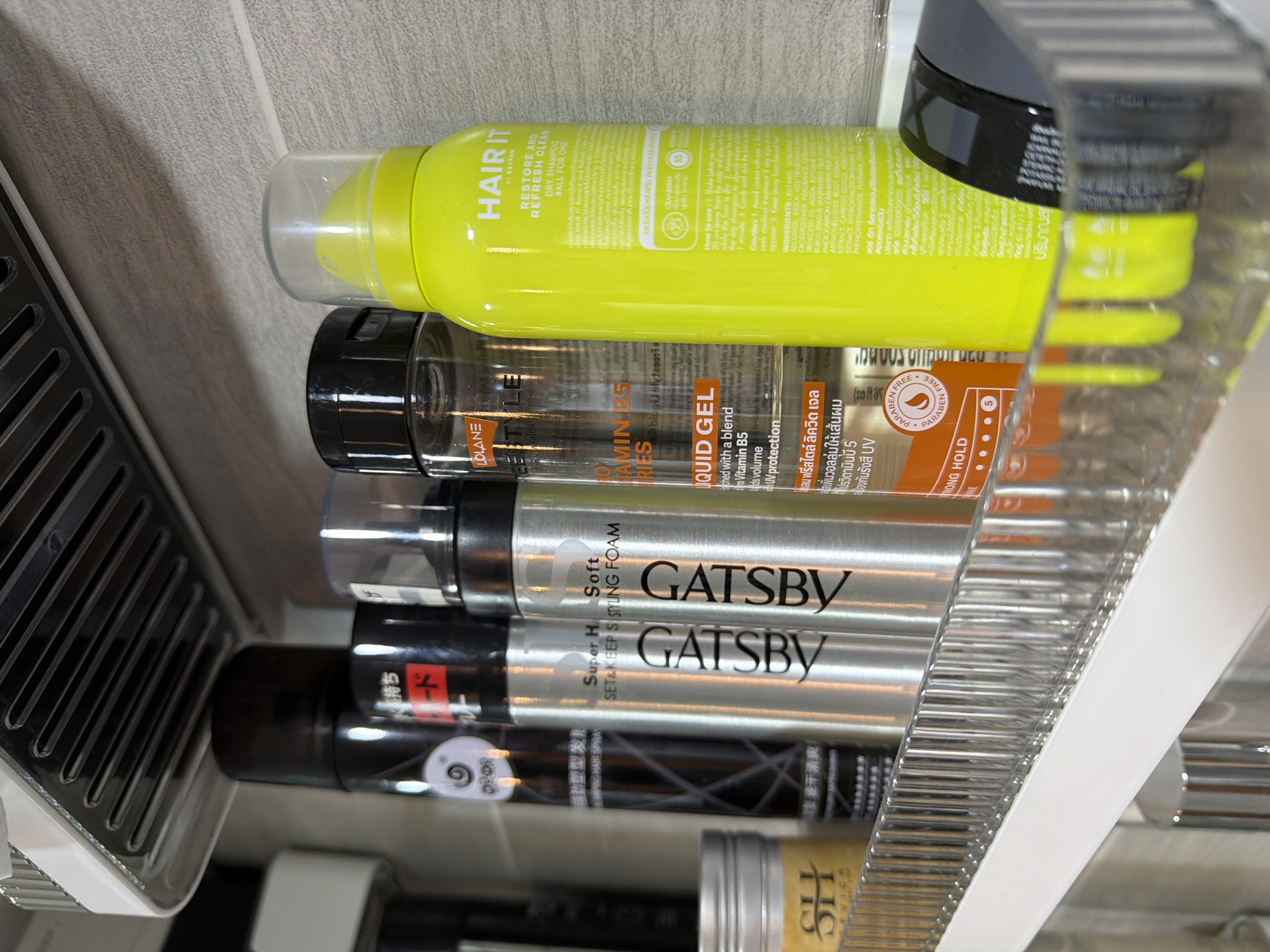 An upward-angled, close-up photograph displays a clear plastic shelf with a white trim, holding an array of hair care and styling products. The shelf is mounted against a light grey tiled wall, characterized by visible horizontal grout lines. A white and clear plastic fixture, possibly a ventilation grate or light, is partially visible at the top of the frame, above the shelf.
From left to right, the objects on the shelf are:
1. A small, silver-colored cylindrical jar with a light yellow label, partially obscured but showing the letters "SH".
2. A tall, black cylindrical can or bottle, partially visible, featuring a white spiral logo and indistinct dark text, possibly Japanese or another East Asian script. Text "HAIR SPRAY" is partially discernible.
3. Two identical silver aerosol cans, both labeled vertically with "GATSBY" in black capital letters. The first GATSBY can specifies "Super Hard SET & KEEP STYLING FOAM". The second GATSBY can specifies "Soft STYLING FOAM".
4. A clear plastic bottle with a black cap, labeled "LOLANE STYLE". Visible text includes "PRO VITAMIN B5 SERIES" and "LIQUID GEL". An orange section on the label notes "PARABEN FREE" and "STRONG HOLD," with a strength rating of "5." Thai script is also present on the label.
5. A prominent neon yellow cylindrical bottle with a clear plastic dispenser cap, labeled "HAIR IT by SAYPAN". The front states "RESTORE AND REFRESH CLEAN" and "DRY SHAMPOO ALL FOR ONE". The reverse side provides detailed instructions, cautions, and ingredients in both English and Thai, noting "CRANBERRY" and "VITAMIN B5" content.
6. A dark, possibly black, circular container with a black lid, likely a hair styling wax or pomade, is partially visible on the rightmost edge of the shelf.
The products are neatly arranged, suggesting storage in a bathroom or personal grooming area. The presence of Thai script on multiple product labels indicates a connection to Thailand. No individuals are present in the image, and no actions or interactions are depicted.
