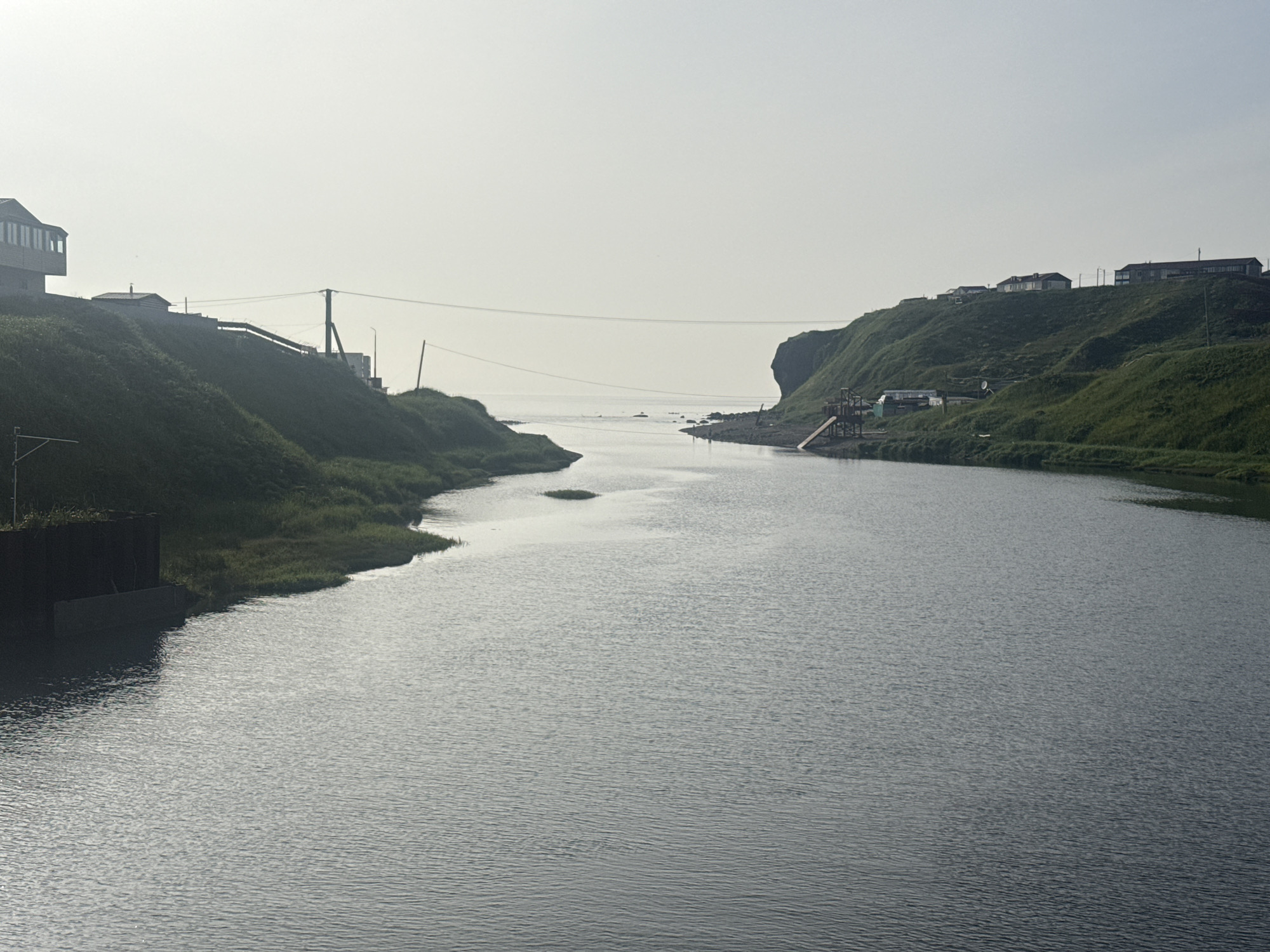 A wide, calm river flows through a grassy valley towards a hazy ocean. On the left bank, a multi-story building with a prominent bay window sits on a hill above a dark, square structure. A transmission tower and power lines stretch across the scene. On the right bank, a grassy cliff slopes down to the water, dotted with several small houses. A wooden structure with a ramp is visible near the water's edge on the right. The sky is overcast and bright, with a faint haze obscuring the distant horizon. The location is Kuril'sk, Russia.