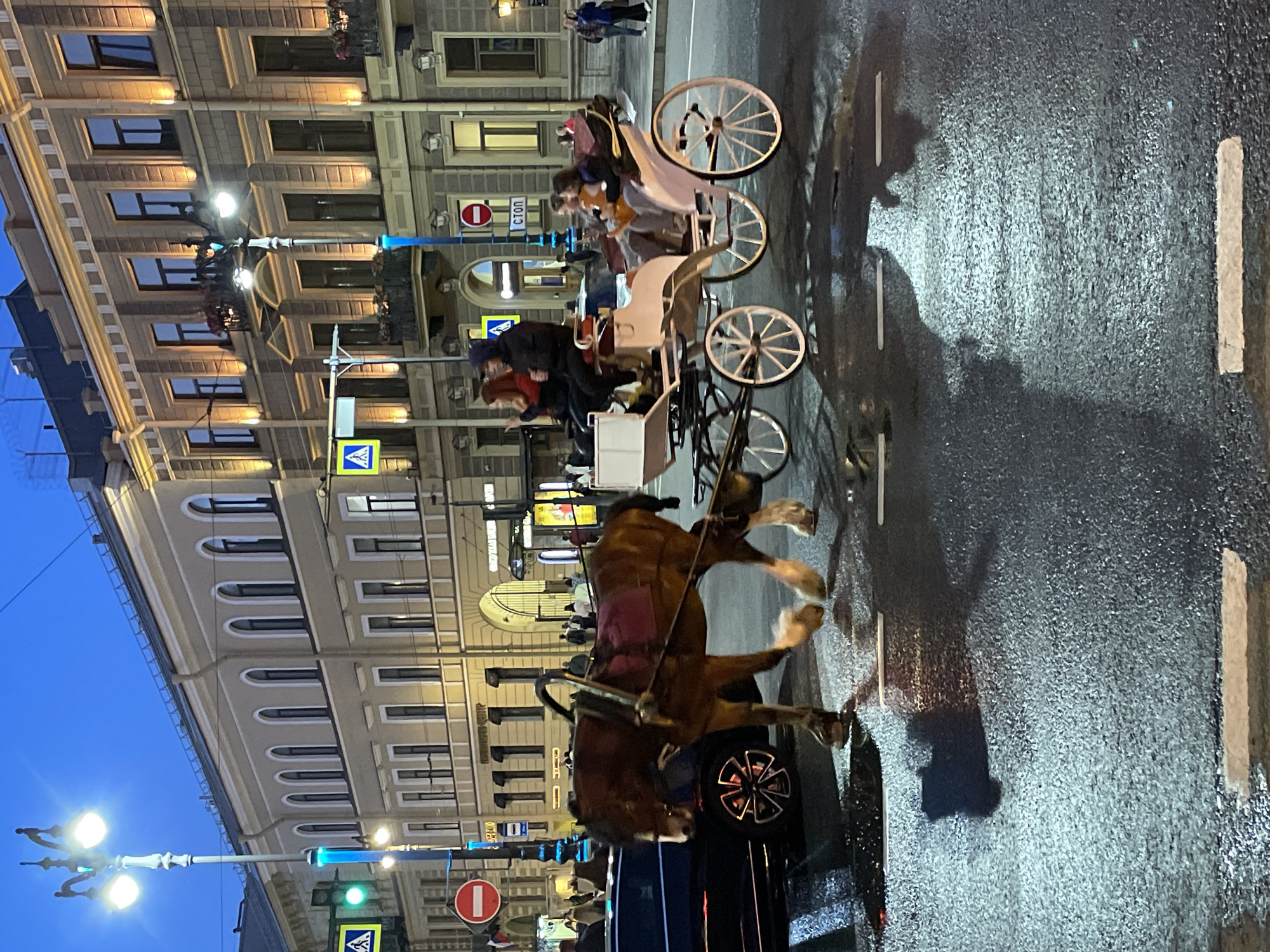 In the city of Unknown, Unknown, a horse-drawn carriage is depicted on a wet city street during twilight. The carriage is light-colored with white spoked wheels and is being pulled by a large brown horse. Several passengers are seated in the carriage. To the left, a dark-colored vehicle is partially visible. The street is wet, reflecting the ambient light from street lamps and building lights. In the background, an ornate building with arched windows and decorative lighting lines the street. Traffic signals and street signs are also visible. The sky is a deep blue, indicating the time of day is likely early evening or late afternoon.