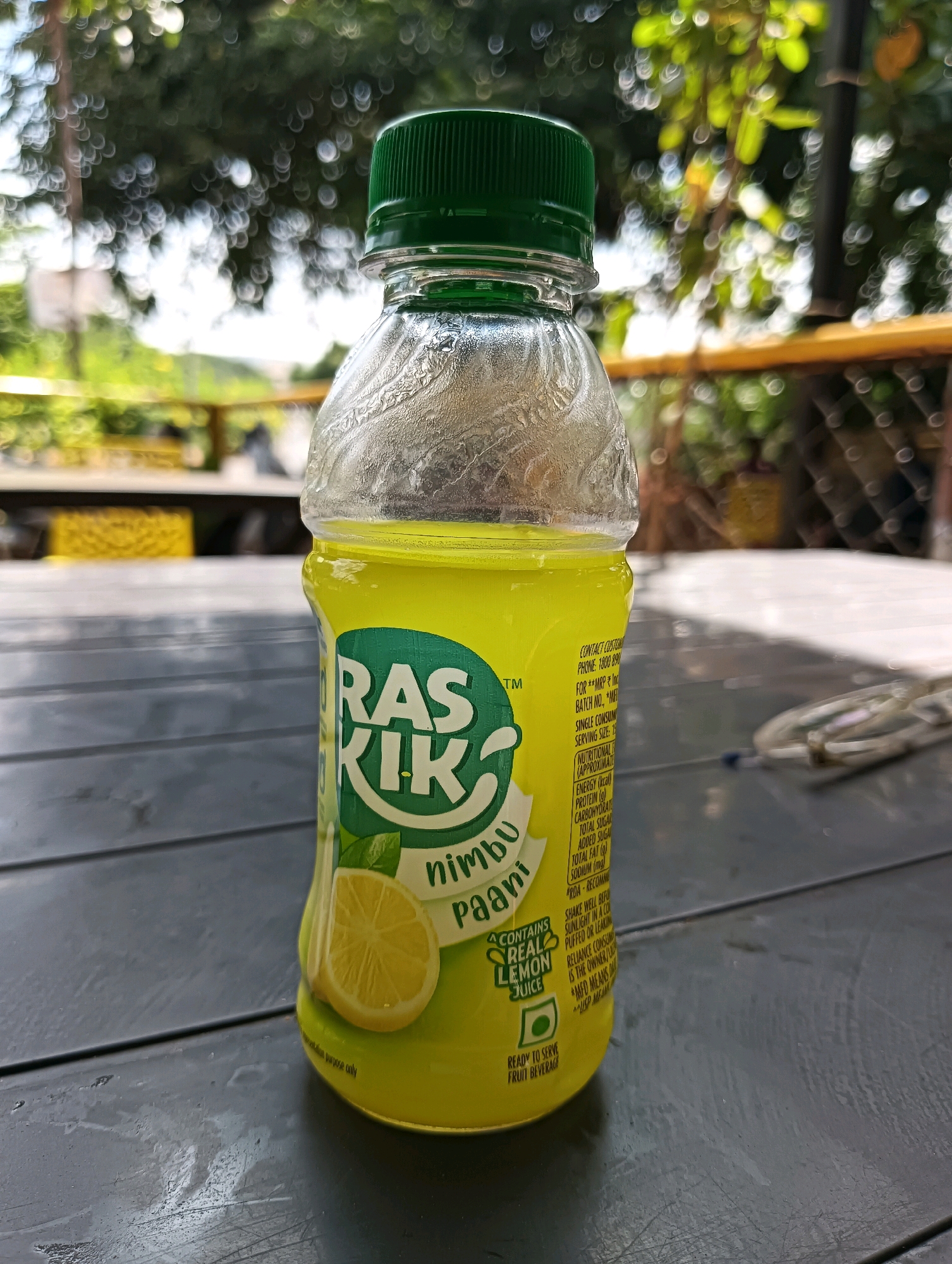A close-up, eye-level shot shows a clear plastic bottle with a green cap and a bright yellow liquid. The bottle is placed on a dark, textured surface, possibly a wooden table. The label on the bottle is white with green text and features a stylized depiction of a lemon slice. Text on the label includes "RAS KIK", "nimbu paani", and "CONTAINS REAL LEMON JUICE". The background is heavily blurred, with indistinct green foliage and streaks of light, suggesting an outdoor setting with dappled sunlight. The overall lighting is bright, creating some glare on the bottle.