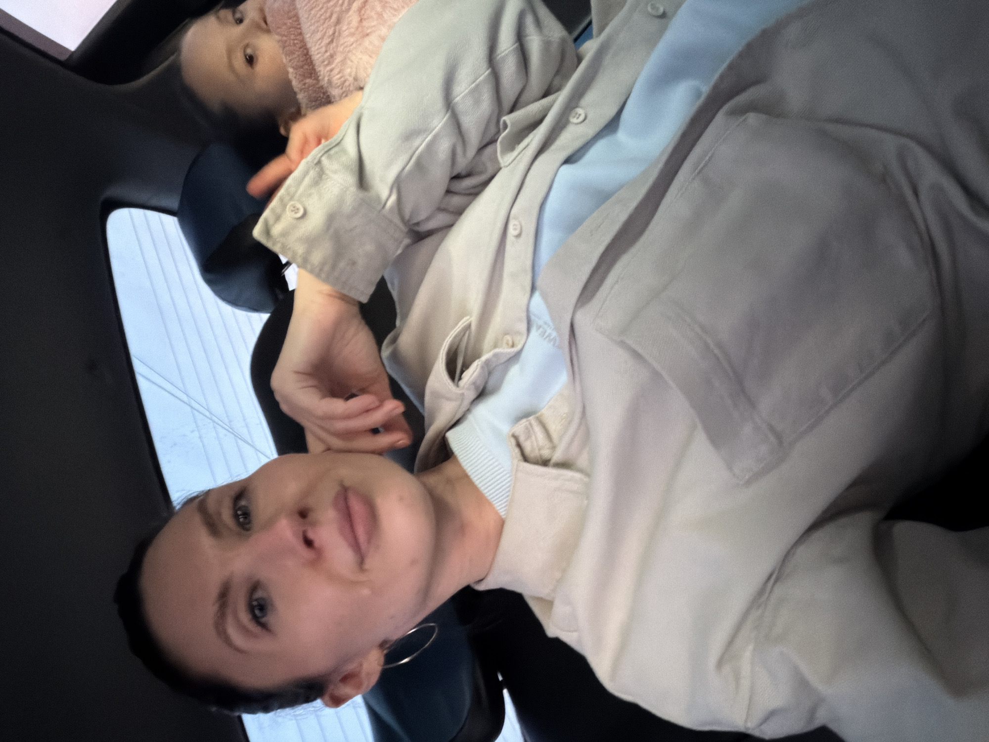 A woman and a child are in a car. The woman is wearing a light-colored button-down shirt over a light blue undershirt. She has dark hair pulled back, blue eyes, and a silver hoop earring. The child, partially visible, is wearing a pink hat. The car interior is mostly black. The background through the window shows a blurry view, possibly indicating movement or a focus on the subjects inside the car. The location, Perm, Russia, cannot be verified from the image.