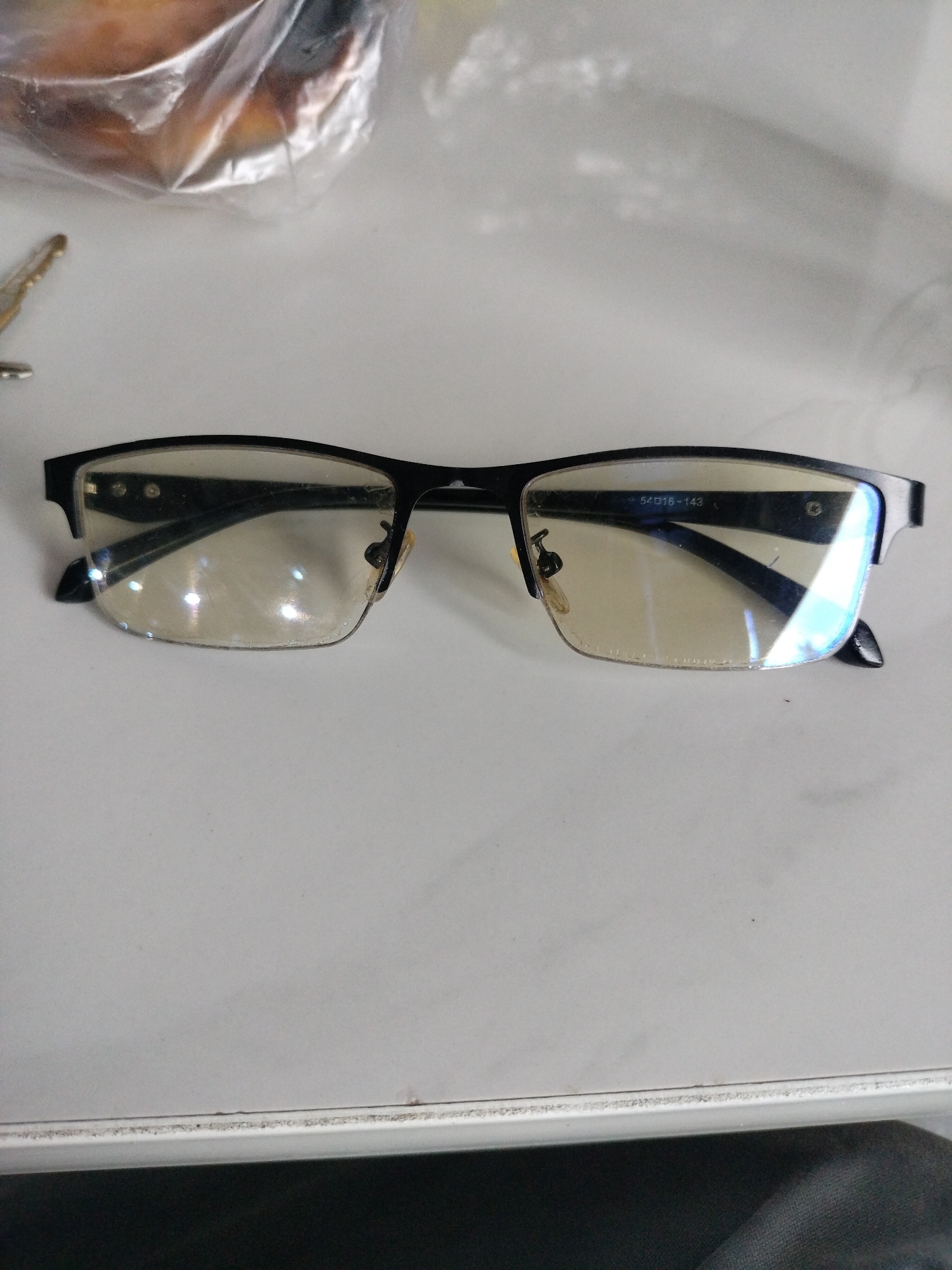 A pair of black-rimmed eyeglasses rests on a white surface. The eyeglasses have semi-rimless lenses that appear to have a yellowish tint. The frame is a matte black metal. On the right temple, markings are visible: "54016-143". A small portion of a plastic-wrapped object and a gold-colored key are visible in the upper left quadrant of the image. The white surface appears to be a tabletop or desk. This scene is not specific to Lhokseumawe, Indonesia.