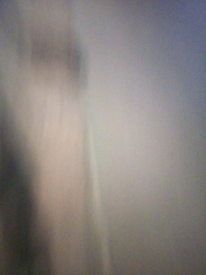 A blurred, indistinct image features a mostly monochrome palette. The left side shows a shadowy vertical form, darker than the overall scene. To the right, the image fades into a lighter, indistinct space with soft, subtle gradients of gray. The focus is absent, making it difficult to identify any specific objects or textures. The effect resembles looking through a fogged lens.