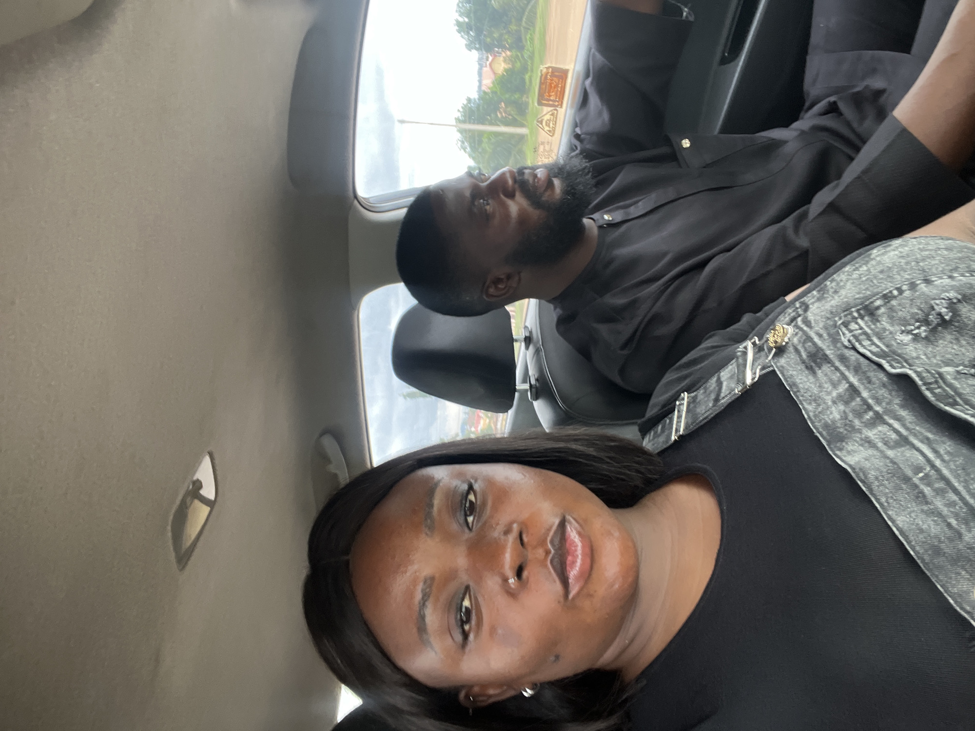 An eye-level, close-up, outdoor selfie shows two Black individuals in the front seats of a car, likely in Galadimawa Village, Nigeria. The woman in the foreground, with a nose ring and hoop earrings, looks directly at the camera with her mouth slightly open. She wears a black top and denim overalls. To her right, the man, with a beard and wearing a black traditional outfit, drives the car and looks to his right. The car's interior, including the ceiling light and headrests, is visible, as is the rural landscape outside the windows, with trees, buildings, and power lines suggesting a Nigerian setting. The lighting is bright and natural, indicating it is daytime.
