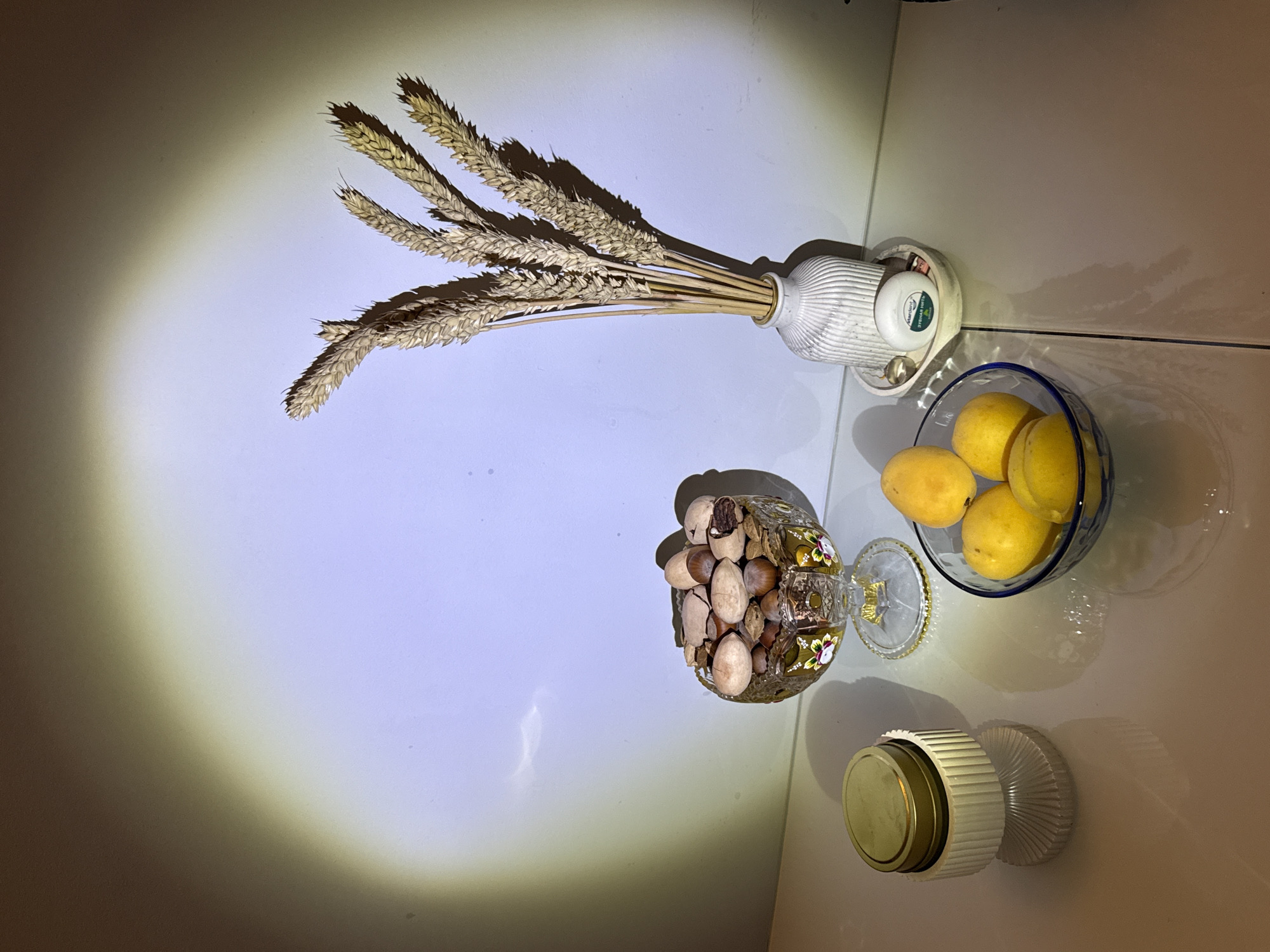 A still life arrangement is displayed on a reflective white surface against a white wall illuminated by a spotlight, casting long shadows. In the center, a decorative glass bowl filled with various nuts sits to the left of a glass bowl containing five yellow fruits, possibly apricots or plums. To the right of the fruit bowl, a white ribbed vase holds a bouquet of dried wheat stalks. A small white dish sits beneath the vase, holding a round green and white dental floss container. To the far left of the image, a two-tiered white object with a gold lid sits on the surface, its reflection visible below. The overall scene is well-lit and composed, with the spotlight creating a dramatic effect.