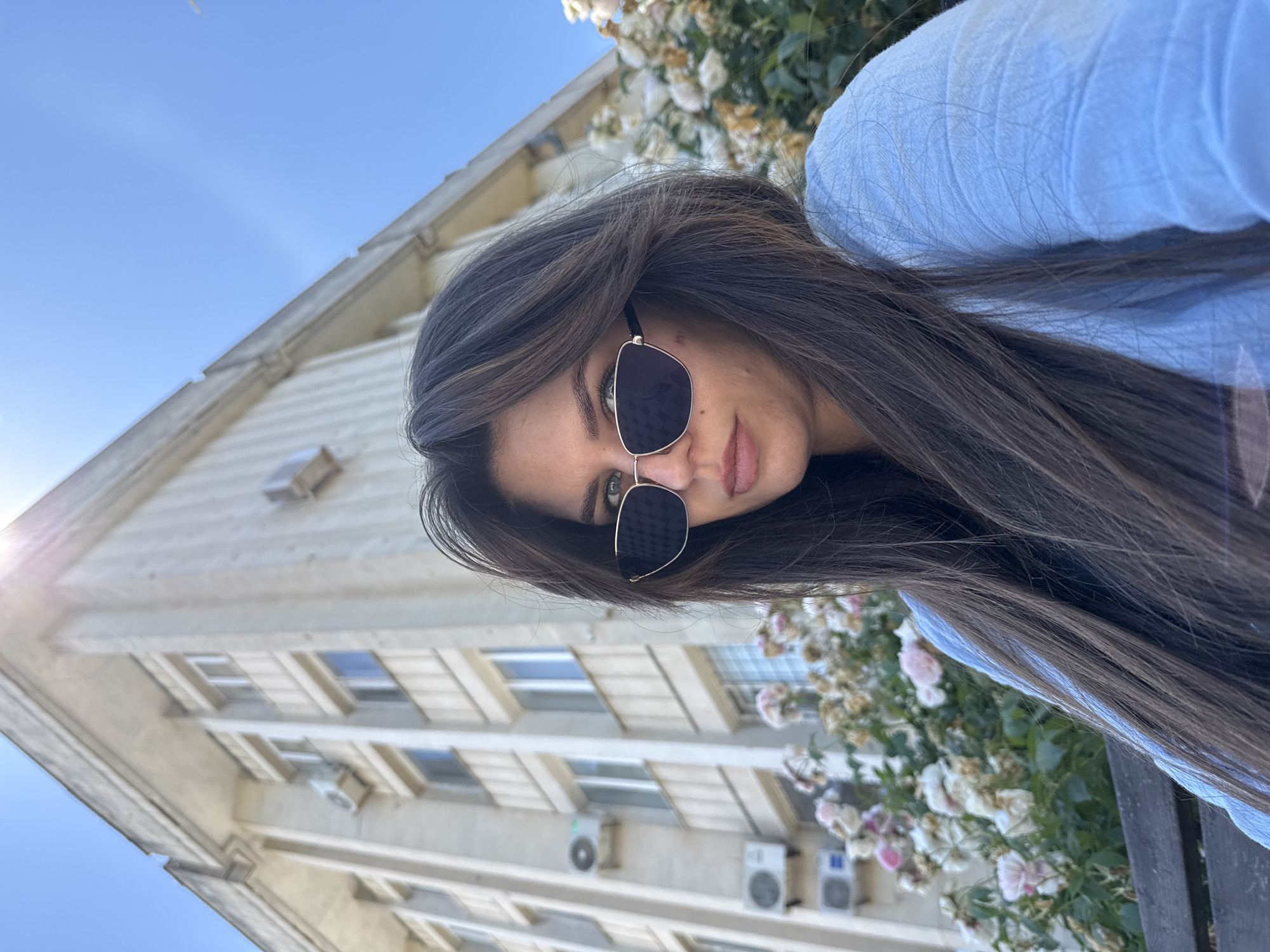 A woman wearing sunglasses and a light blue shirt takes a selfie outdoors. She has long, dark brown hair and is looking towards the camera. Behind her, a beige building with multiple windows and air conditioning units is visible. In the foreground, a bush with white and pink roses is in bloom. The sky is clear and blue.