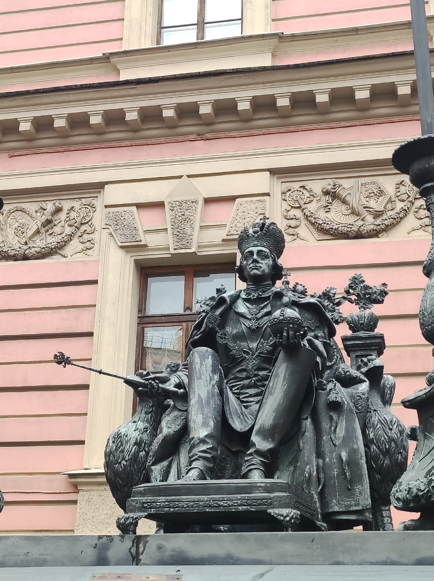 The image shows a bronze statue of Peter the Great, located on Senate Square in Saint Petersburg, Russia. The statue depicts Peter the Great seated on a horse, with his right hand raised and holding a scepter. The horse is rearing up on its hind legs, and the statue is placed on a large granite pedestal. The background of the image shows a pink building with windows and decorative architectural elements. The statue is a prominent landmark and a symbol of Saint Petersburg.