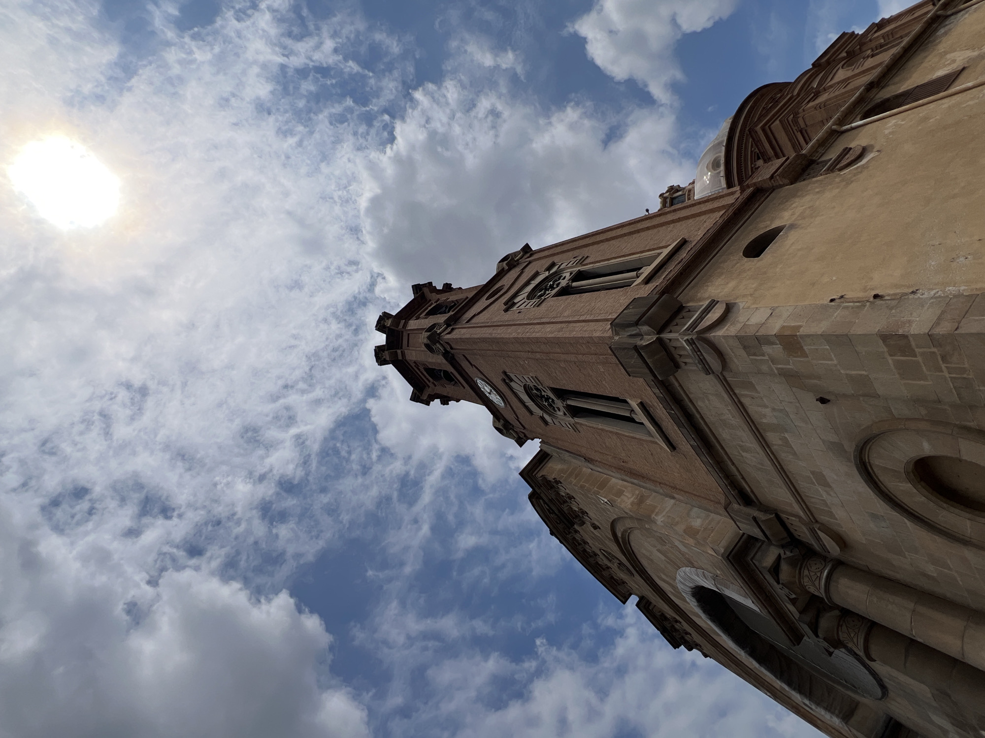 A low-angle shot shows the exterior of a tall, brick church with a domed roof. The building is constructed of red brick and light-colored stone. A clock face is visible on one side of the tower. The sky above is blue with scattered white clouds, and the sun is shining brightly in the upper right corner of the frame, partially obscured by clouds. The architecture suggests a European style, likely a church or cathedral. This is the exterior of the Sagrada Familia church in Barcelona, Spain.