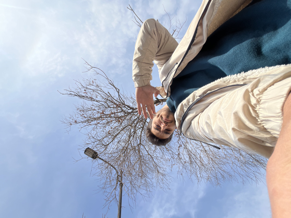 A low-angle selfie depicts a man with a dark-haired head and fair skin. He's wearing a teal sweater and a light beige jacket with black accents. His right hand is slightly raised. In the background, a bare tree with thin branches intersects with a light blue sky dotted with thin clouds. A gray street lamp stands to the left of the tree. The camera angle creates a perspective looking upward, emphasizing the sky and the man's position above the viewer.