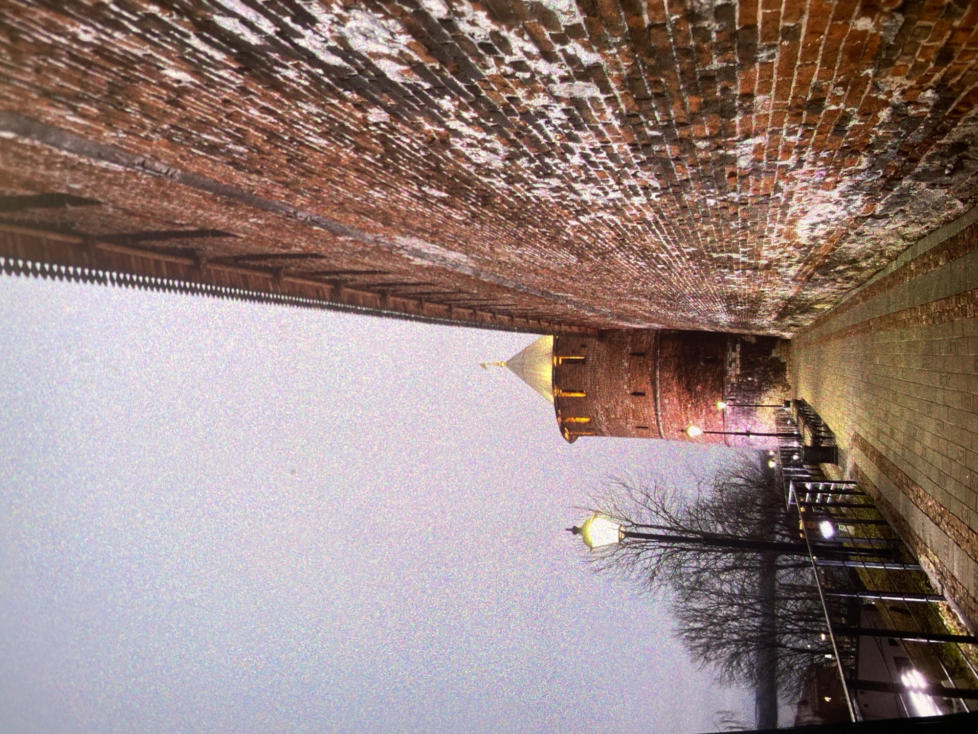 An evening or night scene depicts a historic pathway running alongside a formidable brick wall, leading towards a round brick tower with a conical, possibly gilded or brightly lit, roof in the distance. The image is oriented such that the brick wall is on the left, curving slightly into the background. The paved pathway, illuminated by several streetlights with a warm, yellow-orange glow, occupies the lower right portion of the frame. A dark metal railing lines the right side of the path, separating it from a section with bare-branched trees, indicating a winter or early spring season.
The tower, constructed of red brick, is prominently lit, casting a warm light and highlighting its texture. The sky, a hazy and grainy grey-purple, dominates the upper right, suggesting low light conditions or perhaps a foggy atmosphere. The extensive brick wall shows signs of age and wear, with patches of lighter material, possibly weathered mortar or frost. The overall impression is one of historical architecture illuminated against a muted, twilight backdrop, creating a serene and somewhat mysterious ambiance. No individuals are visible, and there is no discernible text. The image contains significant visual noise, especially in the sky. This scene is set in Saint Petersburg, Russia, likely showcasing a historical fortress or similar monument.