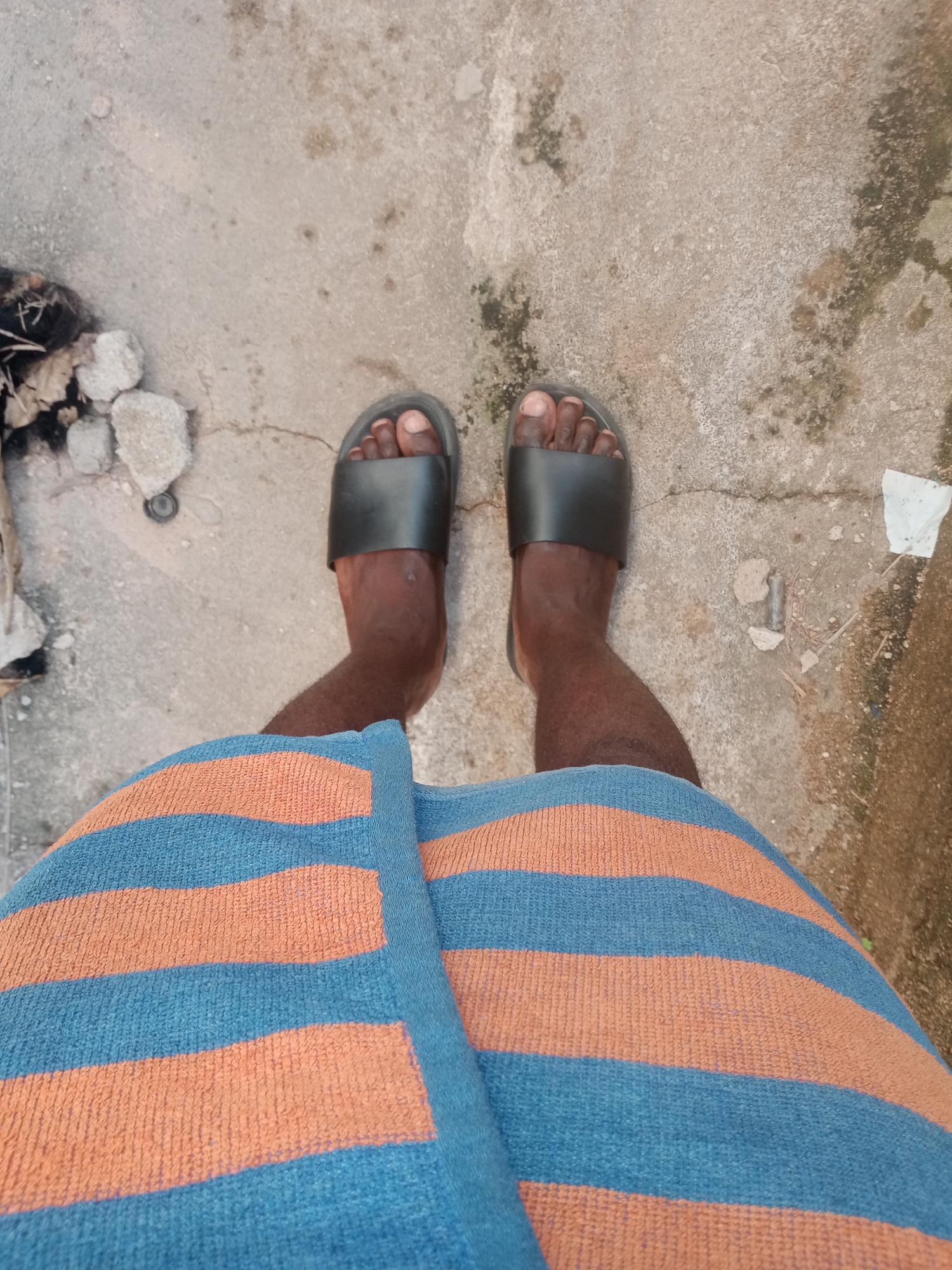 A person's feet wearing black slides are visible, standing on a concrete surface. The person is also wearing a towel with horizontal stripes of orange and blue. The concrete is stained and cracked in places, with some debris scattered around. This scene is likely located on a patio or an outdoor living space in Jalingo, Nigeria.