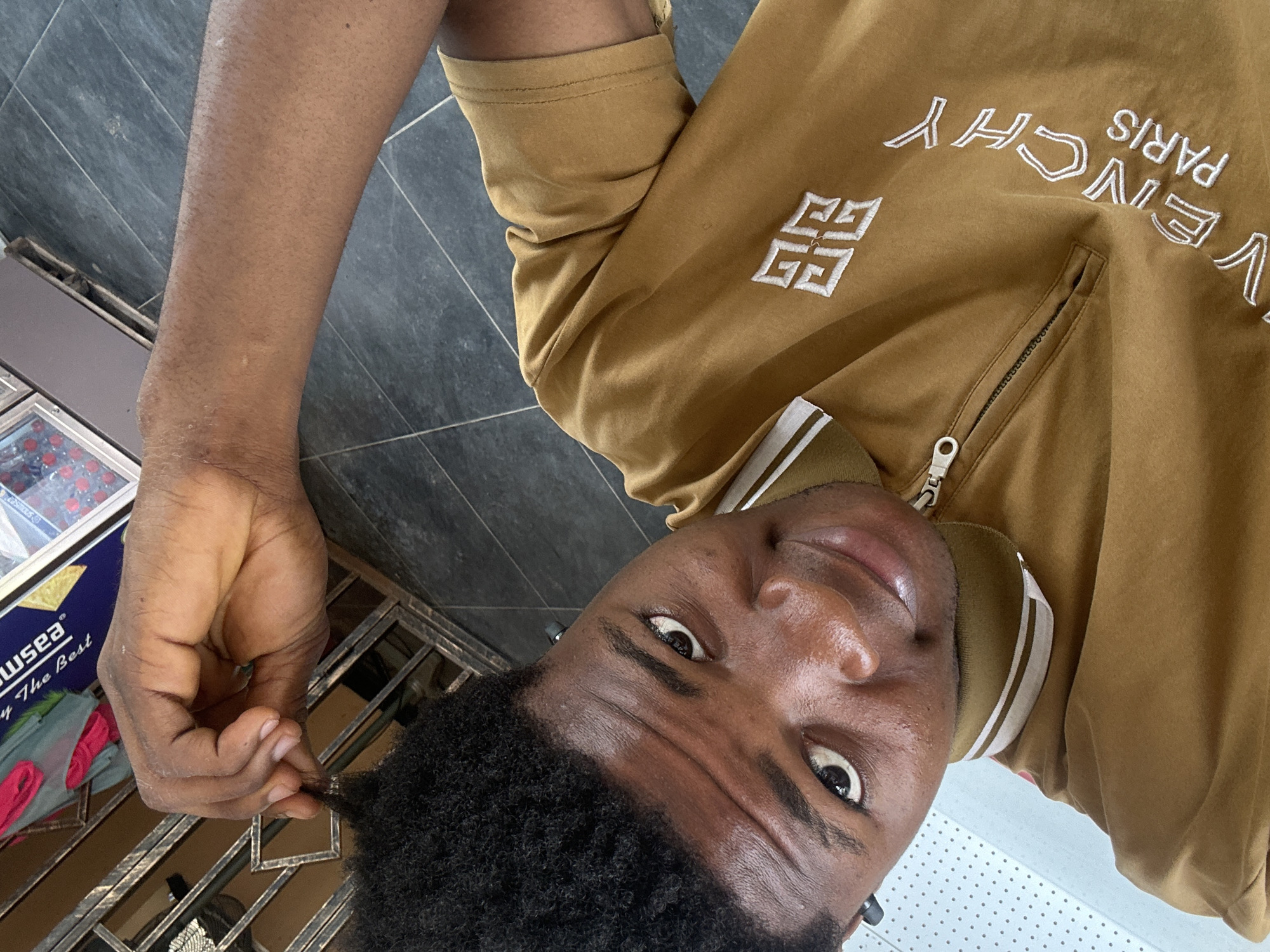 A young Black man is pictured in a selfie taken from a high angle. He has short, dark, curly hair and is looking directly at the camera with a slight smile. He is wearing a brown zip-up polo shirt with white stripes on the collar and "GIVENCHY PARIS" embroidered in white on the front. His right arm is raised, and he is touching the top of his head. The background appears to be an outdoor setting with dark tiled flooring and a metal gate or fence. There are also some blurry elements in the background that suggest a shop or commercial area. The image is taken outdoors in Ihiagwa, Nigeria.