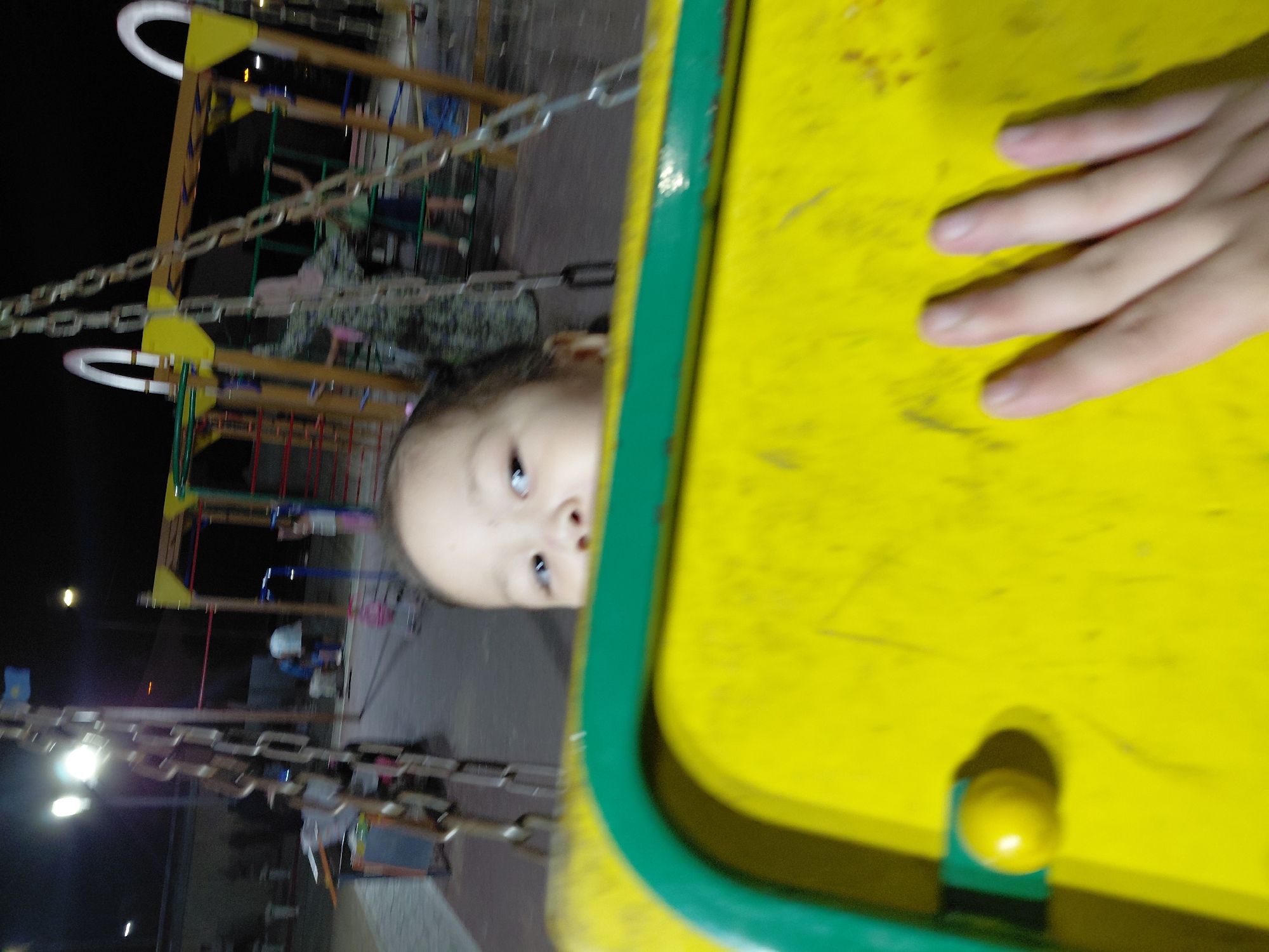 The image shows a close-up of a child's face peeking over a bright yellow surface with a green edge. The child has dark eyes and is looking slightly to the right of the camera. In the background, a playground structure is visible with chains and colorful bars. Several people are blurred in the distance, and the scene appears to be outdoors at night, illuminated by artificial lights. The location is identified as a playground in the city of Aktau, Kazakhstan, suggested by a small blue and yellow flag visible on a pole in the background. A hand with four fingers is also visible in the foreground, resting on the yellow surface. The overall image is somewhat blurry, likely due to the low light conditions and movement.