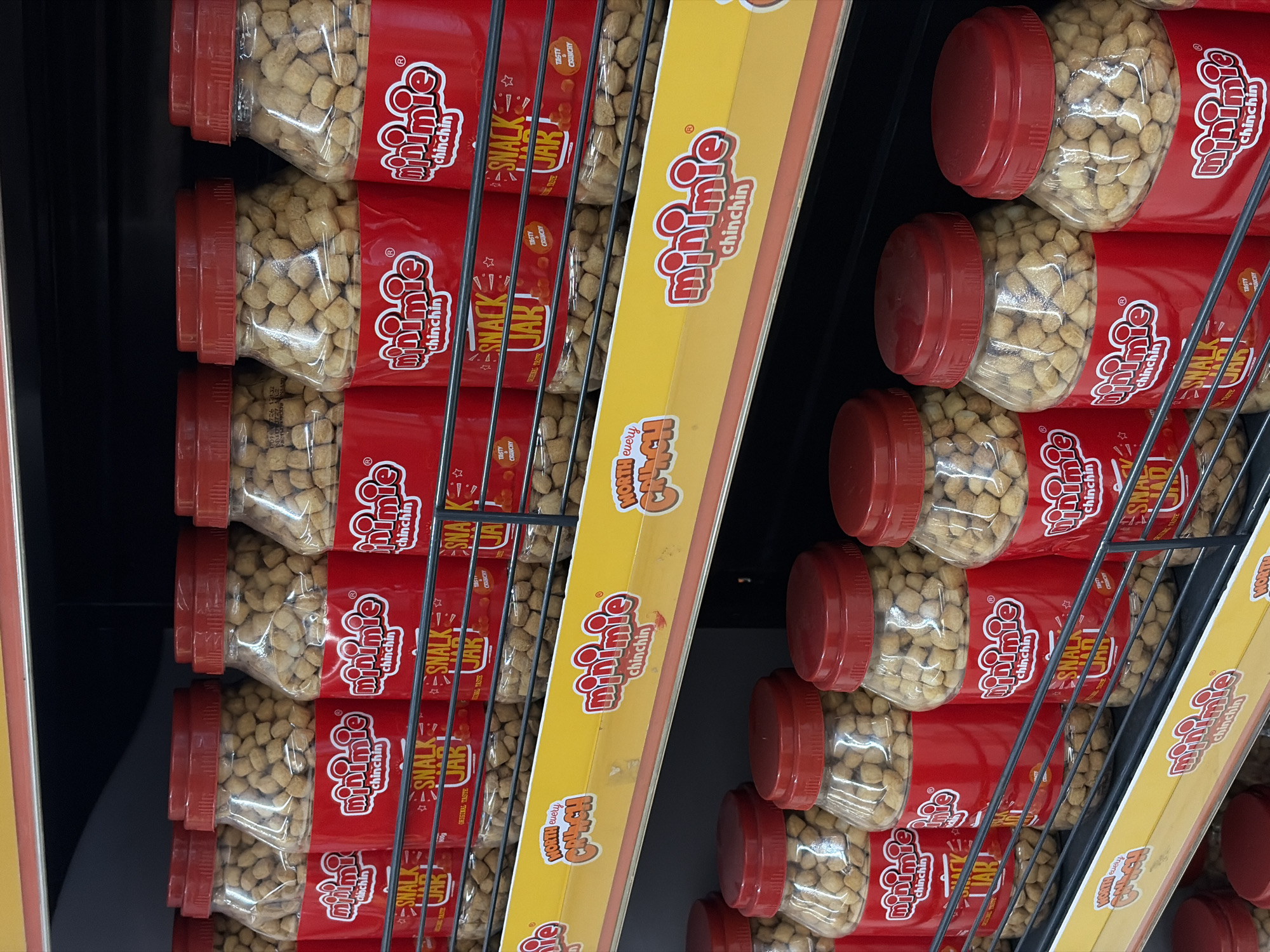The image displays shelves stocked with numerous identical plastic jars of "minimie chinchin" snack, predominantly featuring red labeling and lids. The jars are filled with small, light brown, cubed snacks. The items are arranged on black metal shelving units, suggesting a retail or market environment. A yellow shelf topper with the "minimie chinchin" logo is visible. The overall impression is one of commercial product display in a store. The specific location, Igboefon, Nigeria, is not visually identifiable from the image content itself.