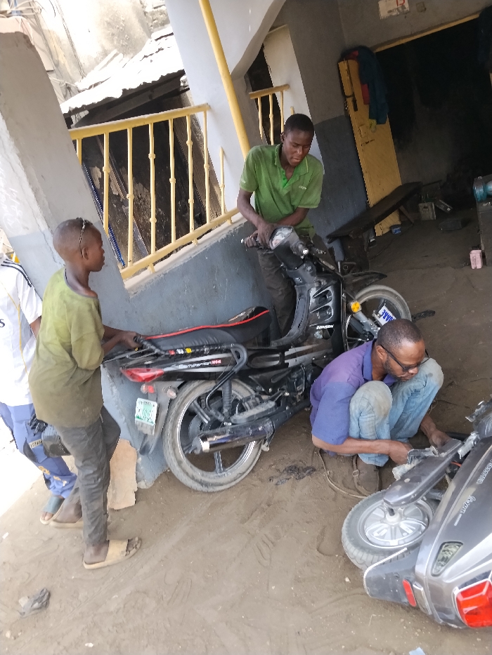 In Fagge D2, Nigeria, an outdoor scene depicts four males engaged in motorcycle and scooter maintenance on a dirt ground surface. To the left, a partial view of a male in a white and blue shirt is visible. Center-left, a younger male wearing a dirty olive-green t-shirt and dark trousers, with light-colored slides, holds the rear rack of an upright black Bonbon 125 motorcycle. The motorcycle displays a Kano license plate. A third male, positioned centrally in a green polo shirt and dark trousers, grips the handlebars of the black motorcycle. To the right and in the foreground, a fourth male, wearing a blue polo shirt, jeans, and brown shoes, crouches while actively working on a grey scooter lying on its side. The background features a building structure with grey and white walls, yellow vertical support beams, and a railing. A dark interior opening of the building shows indistinct objects.