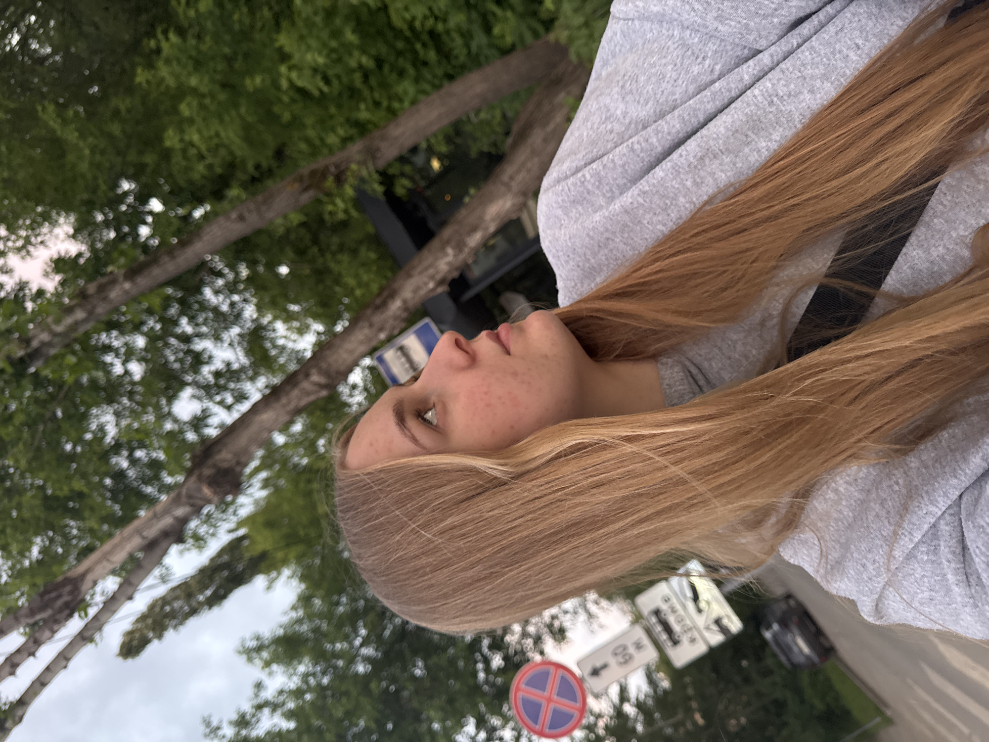 A photograph shows a young woman with long blonde hair in profile, wearing a gray hooded sweatshirt. She is outdoors, likely on a street in the city of Perm, Russia. Street signs in the background indicate local traffic regulations and speed limits (60 km/h), as well as parking restrictions (red and blue X on a white circle). Trees are visible in the background. The sky is overcast.