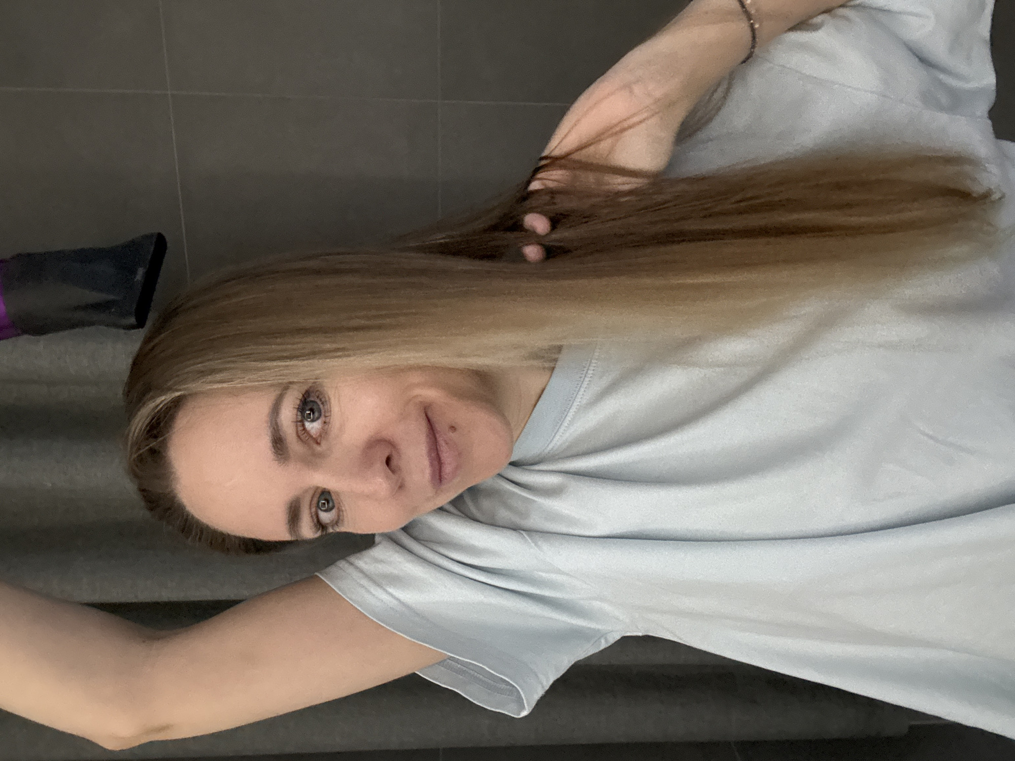 A woman with long, light brown hair is looking at the camera and smiling slightly. She is holding a section of her hair in her hand. A black hair dryer is visible in the background. The woman is wearing a light blue t-shirt and a silver bracelet. The background appears to be a gray tiled surface. The city of Chaikovsky, Russia is not discernible from the image.