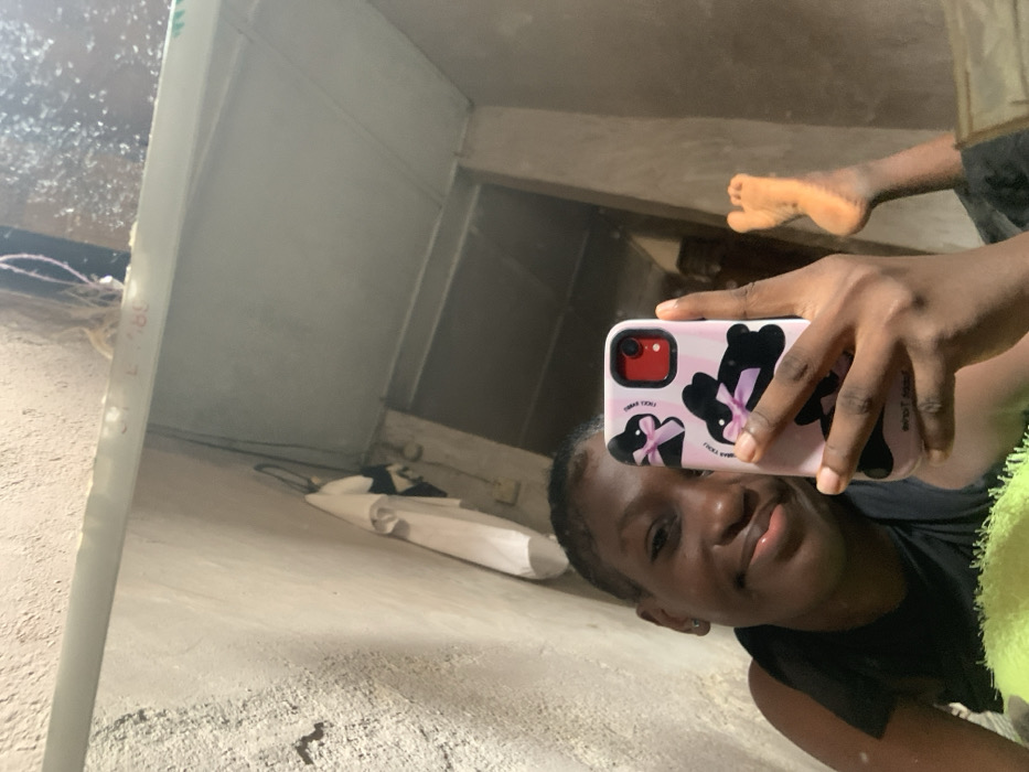 A forensic analysis of an interior scene, observed through a mirror selfie, presumably within a residence in Agege, Nigeria. The primary subject is an individual, appearing female and of African descent, positioned in a reclining manner, smiling directly into the phone's lens. They wear a dark sleeveless top. The subject holds a light pink smartphone, likely an iPhone given the distinct camera bump, encased in a cover featuring multiple black rabbit figures with pink bows, and the phrase 'FUCK YOU' visible in dark lettering. The phone's camera module has a prominent red border. The subject's bare right foot, with toes flexed upwards, is visible in the lower right quadrant of the mirror's reflection. The mirrored environment reveals rough, unpainted concrete walls with a light tan or grey hue. In the upper left, a white shirt and a dark piece of clothing hang from a hook. Exposed electrical wiring is present near the top edge. A dark rectangular opening, possibly a doorway, is visible in the background, leading to an adjacent area. A light-colored structural beam or shelf runs horizontally across the upper portion of the mirror, displaying faint red markings such as 'L18B'.