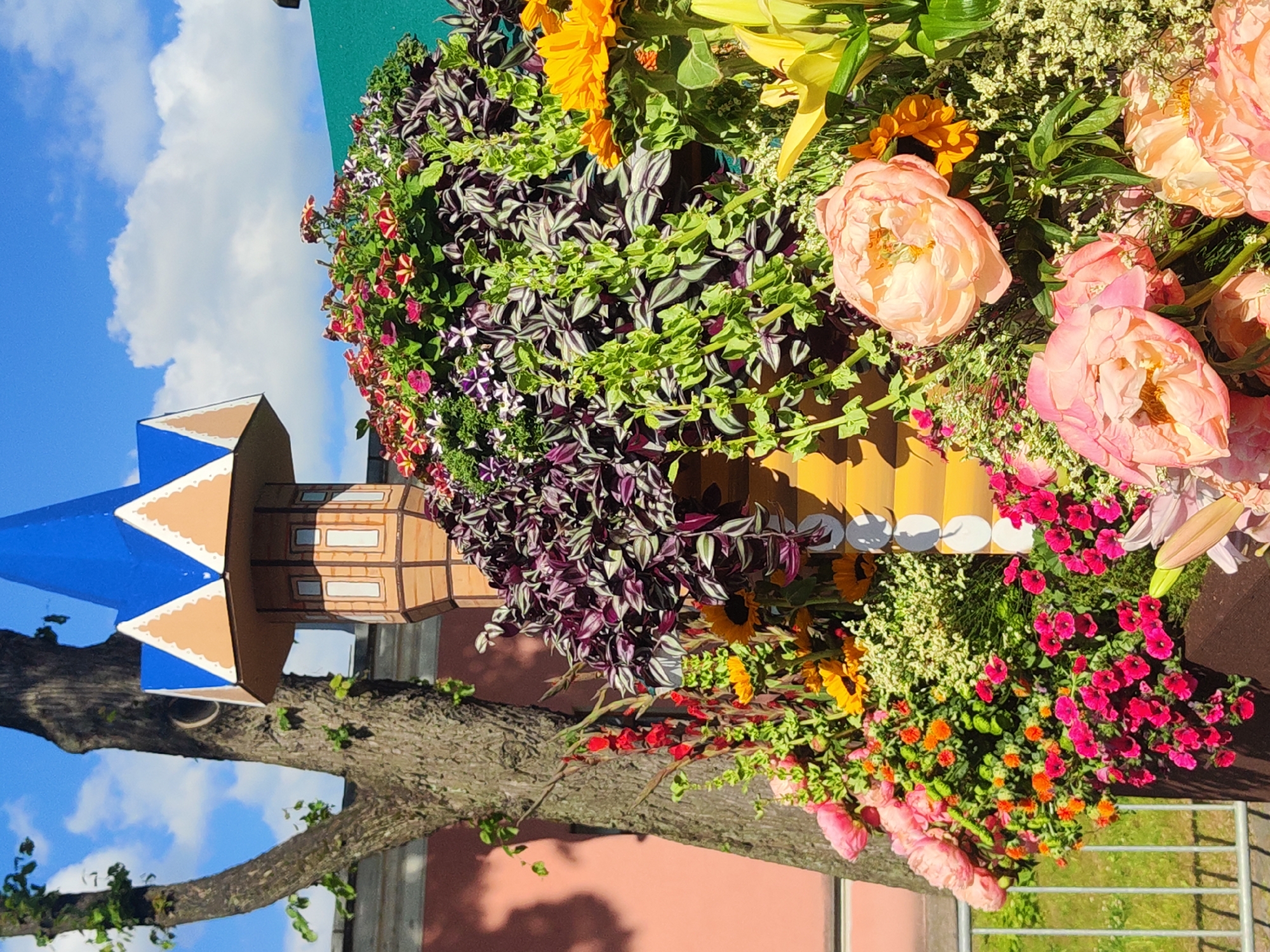 The image depicts a vibrant floral arrangement in front of a building with a pink facade and a green roof. The arrangement includes a variety of flowers such as pink peonies, orange calendula, red gladioli, yellow lilies, and numerous smaller pink and purple flowers. A decorative tower-like structure with a blue roof, resembling a fairytale castle turret, is partially visible behind the flowers. A tree trunk with textured bark stands to the left of the tower. The scene is set outdoors, under a blue sky with scattered white clouds. A metal fence is visible in the foreground. The background suggests an urban environment, possibly a park or public space within Saint Petersburg, Russia, given the architectural style of the turret, which is reminiscent of Russian traditional architecture. No people are present in the image.