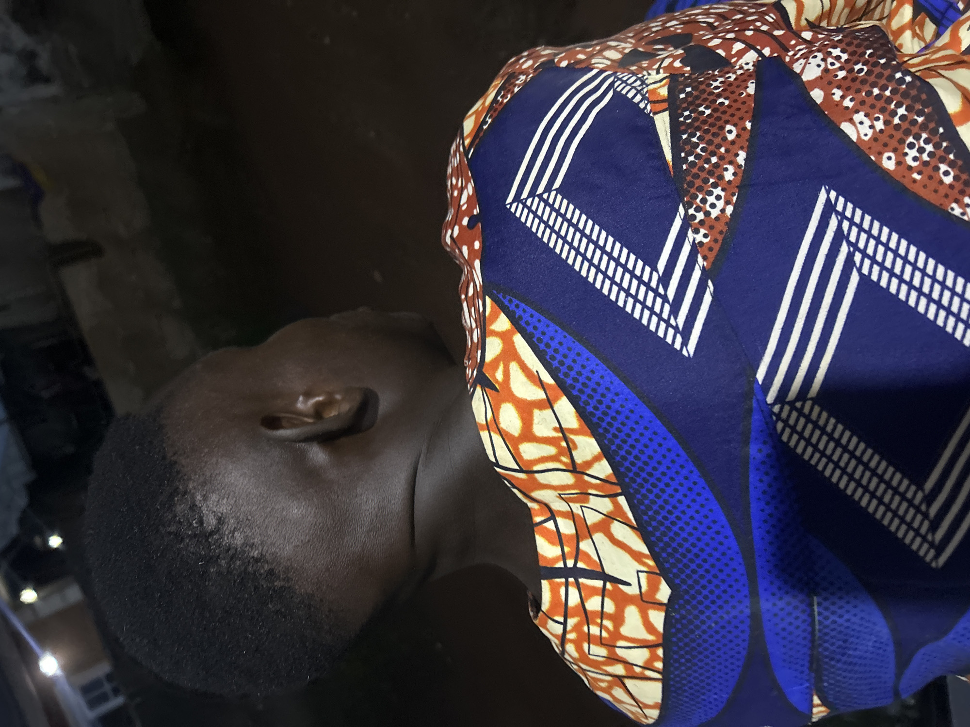 The image shows a person from the back, facing away from the camera, wearing a colorful, patterned shirt. The shirt features geometric designs in blue, white, orange, and red, with some areas appearing to have a dotted or speckled texture. The person has dark skin and short, dark hair. The background is dark and out of focus, suggesting an outdoor setting at night. Hints of artificial lights are visible in the upper left portion of the image, possibly indicating streetlights or lights from buildings. The image quality is somewhat grainy, consistent with low-light photography. The scene is located in Ikorodu, Nigeria.