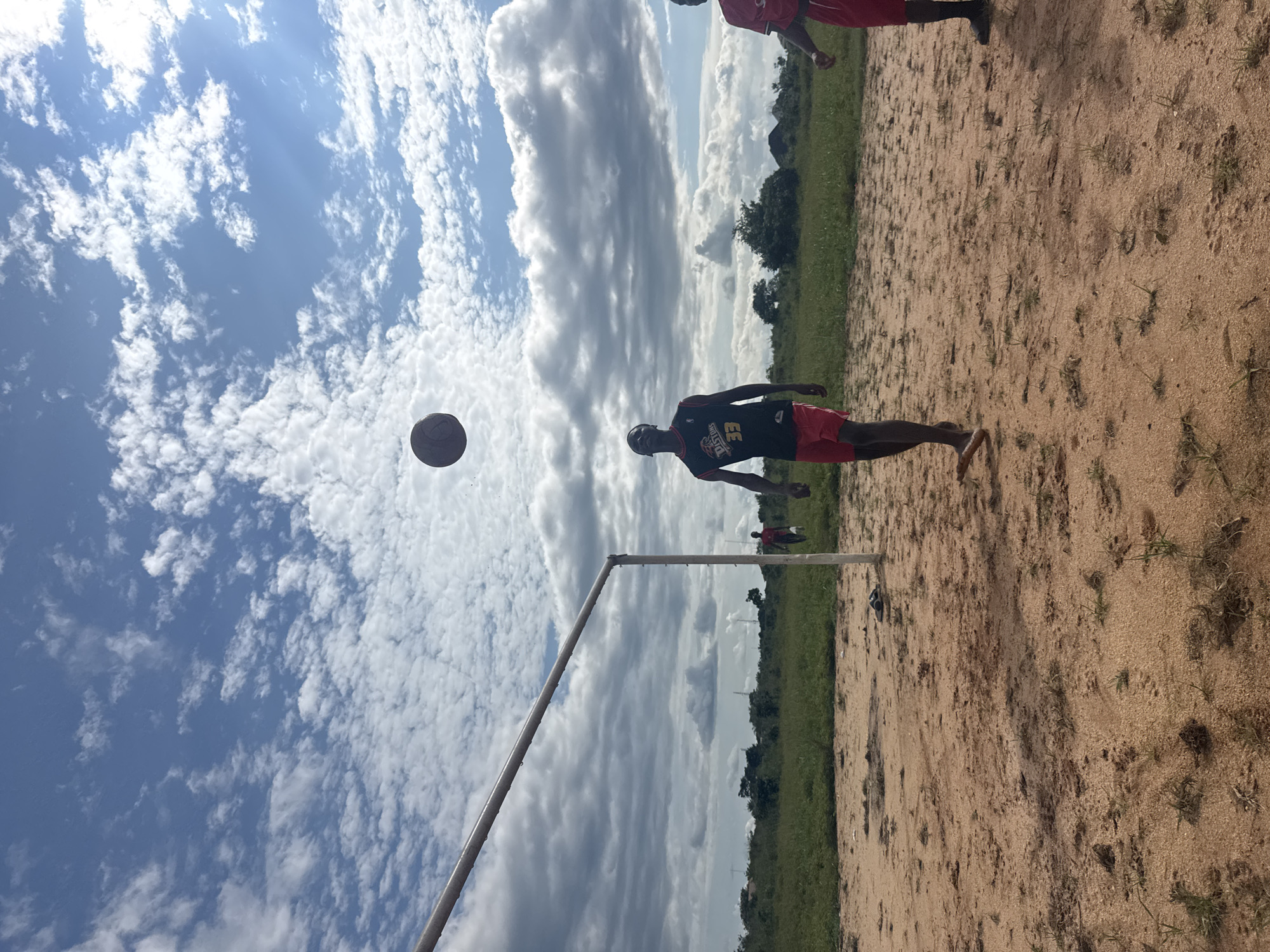 In Ihiagwa, Nigeria, on a dusty, outdoor field, two young men are engaged in a game of soccer. The main subject, a young man wearing a black and red basketball jersey with the number 33 and red shorts, stands near a soccer goalpost, his eyes focused on a soccer ball suspended in mid-air above him. To his right, another young man in a red shirt and black shorts is partially visible, also seemingly participating in the game. The environment is characterized by dry, sandy ground with sparse patches of green grass. In the background, a line of trees and vegetation borders the field, with a hazy sky filled with scattered white clouds above. The overall scene depicts a casual soccer match in a rural or semi-urban setting in Nigeria.
