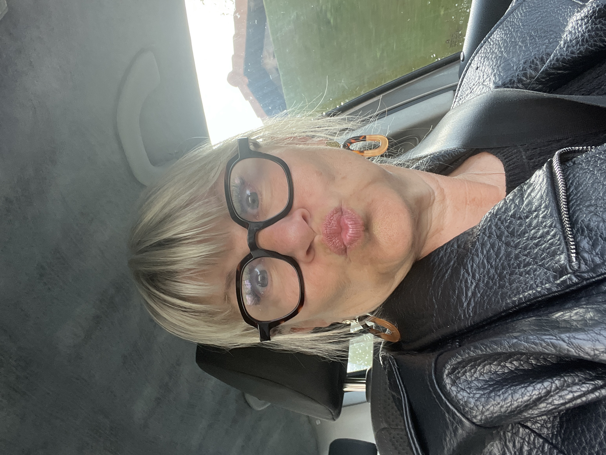 A mature woman with blonde hair and black-rimmed glasses is taking a selfie inside a car. She is wearing a black leather jacket and a seatbelt is fastened across her chest. Her lips are pursed. The interior of the car is visible, including a headrest and a grab handle. A glimpse of the outside environment through the car window suggests a partly cloudy sky. The location, Saint-Bonnet-les-Oules, France, cannot be definitively determined from the image.