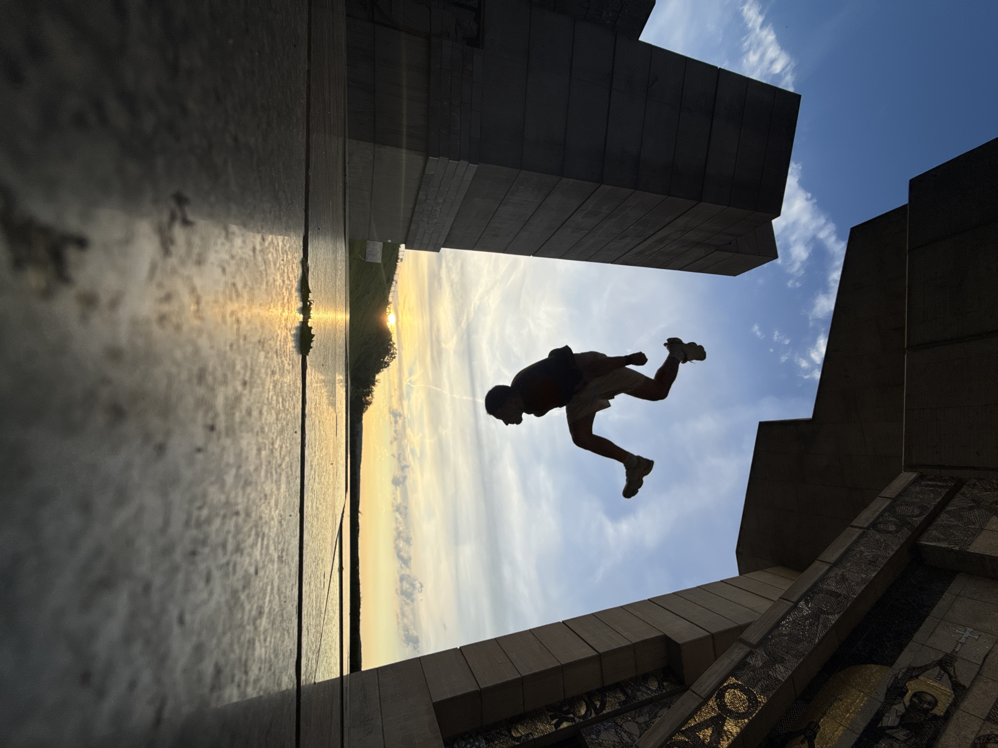 In Shumen, Bulgaria, at the Monument to the Declared Bulgarian State, a silhouette of a person is captured mid-flip against a vibrant sunset sky. The individual, wearing shorts and sneakers, is suspended in the air, performing an acrobatic maneuver. The foreground is dominated by the polished stone of the monument's plaza, reflecting the warm hues of the setting sun. Two imposing, dark stone structures flank the central space. To the left, a monolithic, rectangular pillar rises, its surface textured with horizontal lines. To the right, an ornate section of the monument features geometric patterns and mosaic details, framed by a large, angled concrete beam. Beyond the plaza, a body of water stretches towards the horizon, where the sun dips low, casting a golden glow on the landscape and creating a mirror-like surface. The sky is a gradient of blue, transitioning to yellow and orange near the horizon, with scattered wispy clouds. The overall scene is dynamic, with the action of the flip contrasting with the static, monumental architecture and the serene natural beauty of the sunset.
