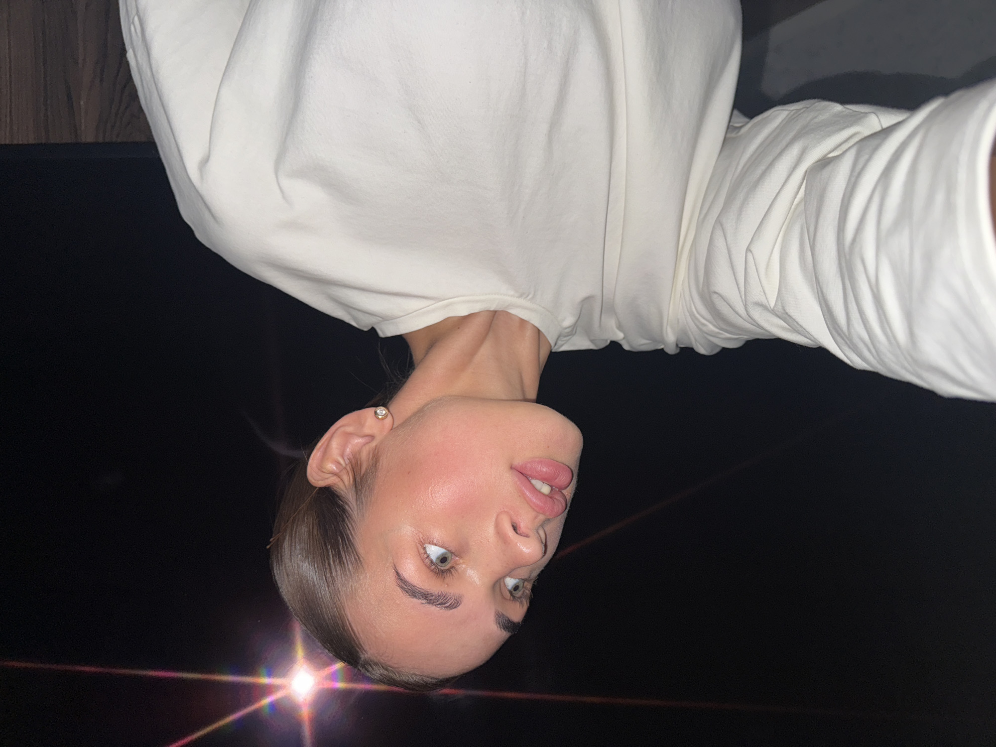A portrait of a fair-skinned young woman with hazel eyes and brunette hair pulled back, wearing a white long-sleeved shirt, is set against a dark background with a bright, refracting light source at the bottom of the frame. The photograph is oriented upside down. The location, Лобаново, Russia, is not visually discernible from the image.