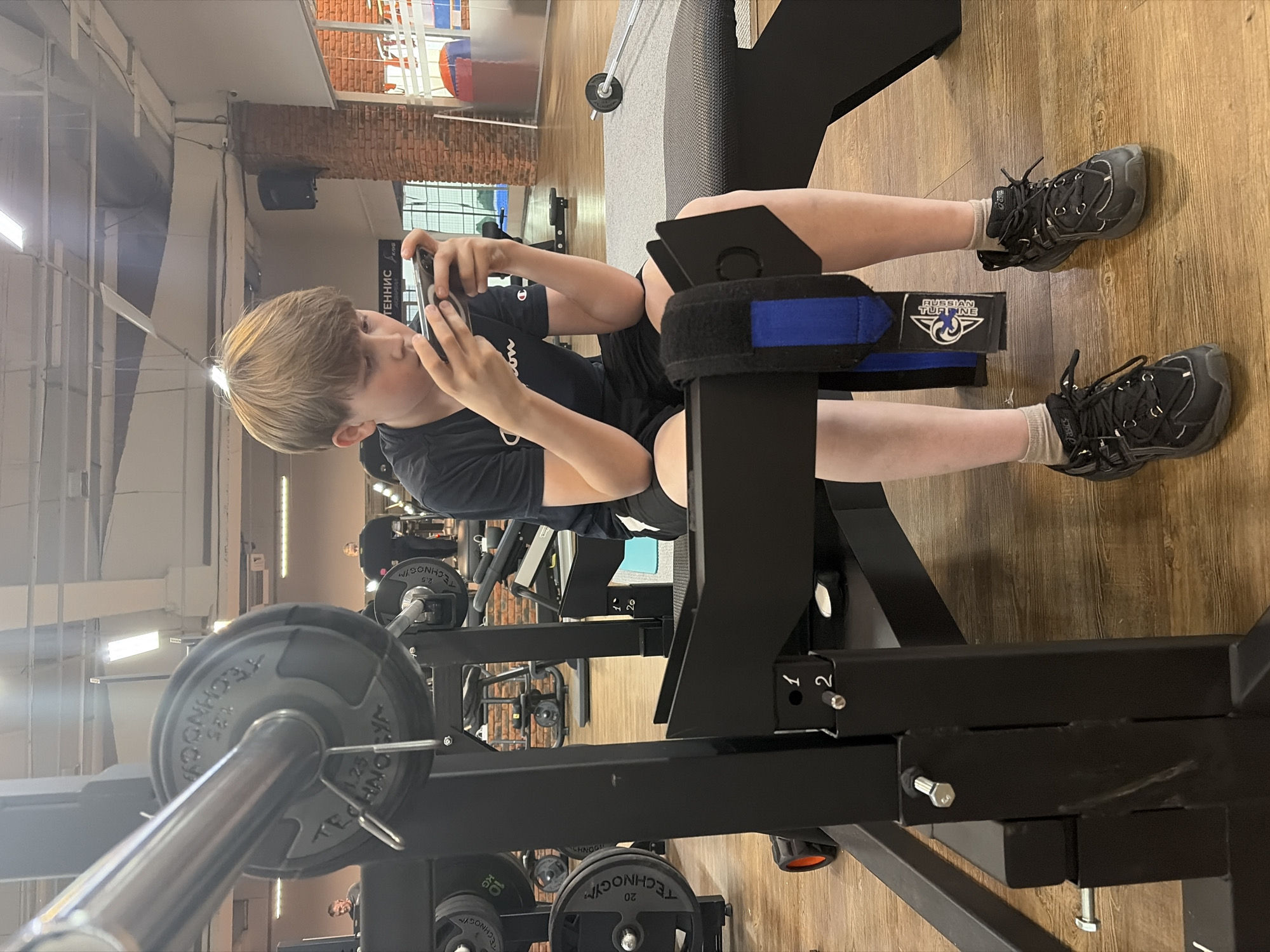 An indoor full-shot image captures a young individual, likely a male, seated on a specialized leg training machine within a gym or fitness center. The person has blonde hair and is wearing a black t-shirt, black shorts, white socks, and black sneakers. They are holding and looking intently at a smartphone with both hands, appearing to be taking a break or waiting.
The gym features a wooden floor and walls with a combination of light-colored panels and exposed red brick. Bright overhead lighting illuminates the space. Various pieces of black metal gym equipment are visible, including barbells and weight plates from the "TECHNOGYM" brand, with markings like "10 KG", "20", and "2.5". One of the person's legs is secured by a blue and black strap with the text "RUSSIAN TURBINE" and a logo featuring a stylized 'R'. The sneakers worn are "Asics" brand, partially visible as "Jasics" in the OCR. In the background, a sign displays "ТЕННИС", which translates to "Tennis," suggesting the facility might include tennis courts. Adjustment pins on the equipment are marked with "1" and "2". The scene provides no direct indication of the time of day, but the artificial lighting suggests it could be any time. The location is Chaikovsky, Russia, aligning with the Russian text and brand name.