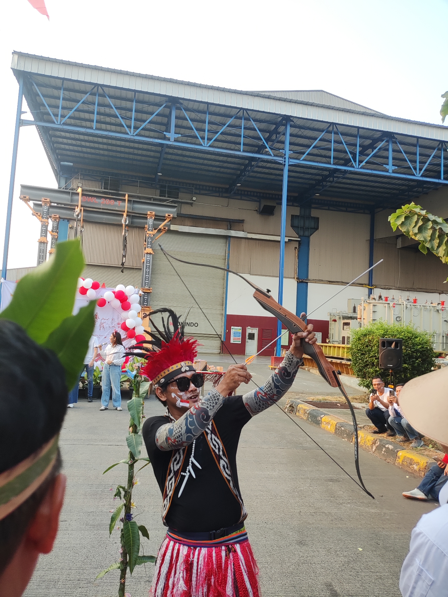 In East Jakarta, Indonesia, an archer adorned in traditional attire, featuring a red headdress with feathers, face paint, and a patterned vest, holds a bow and arrow. The archer is aiming the arrow, with the bowstring drawn back. Behind the archer, a group of spectators are gathered. In the background, there is a large industrial building with a corrugated metal roof and blue metal framework. Red and white balloons are visible, along with some trees and electrical transformers. The scene appears to be an outdoor event, possibly a cultural festival or performance, taking place in an industrial area.