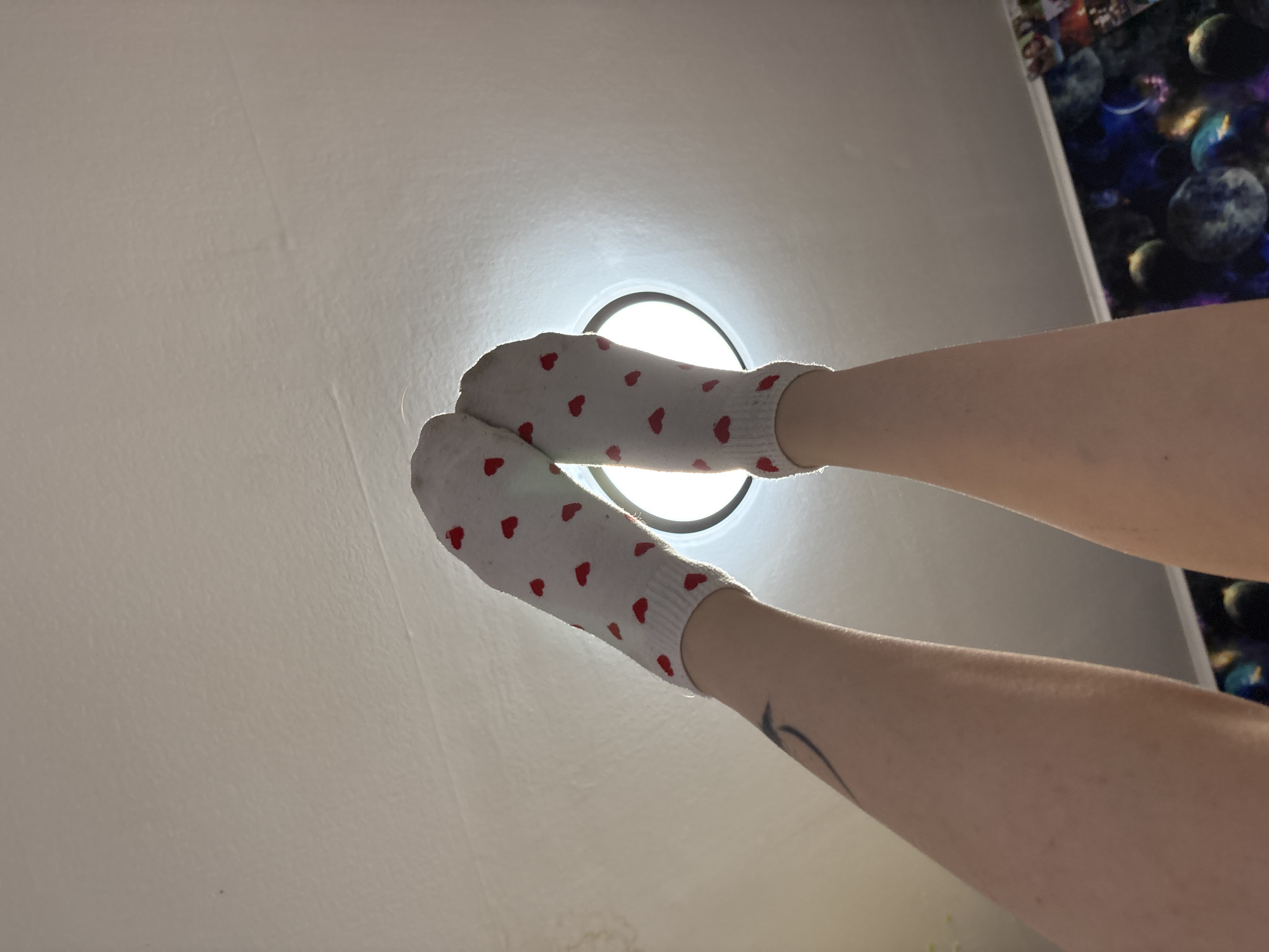 The image shows a pair of feet wearing white socks with red hearts, positioned against a white ceiling with a recessed light fixture. One leg has a tattoo on the lower calf. Part of a wall with space-themed decor is visible in the upper right corner.
The location is not discernible from the image.