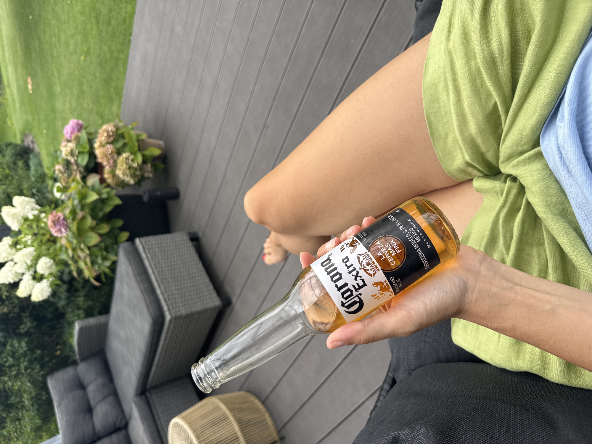 A person's hand holds a partially consumed bottle of Corona Extra beer, its amber liquid visible. The person is seated on a dark gray outdoor sofa, their legs draped in a light green fabric and partially visible, with a sliver of a blue garment beneath. The scene unfolds on a gray composite deck, surrounded by lush green grass and potted hydrangeas in shades of white, pink, and muted brown. In the background, another piece of outdoor furniture, a light-colored wicker side table, is visible. The location is identified as Carnikava, Latvia.