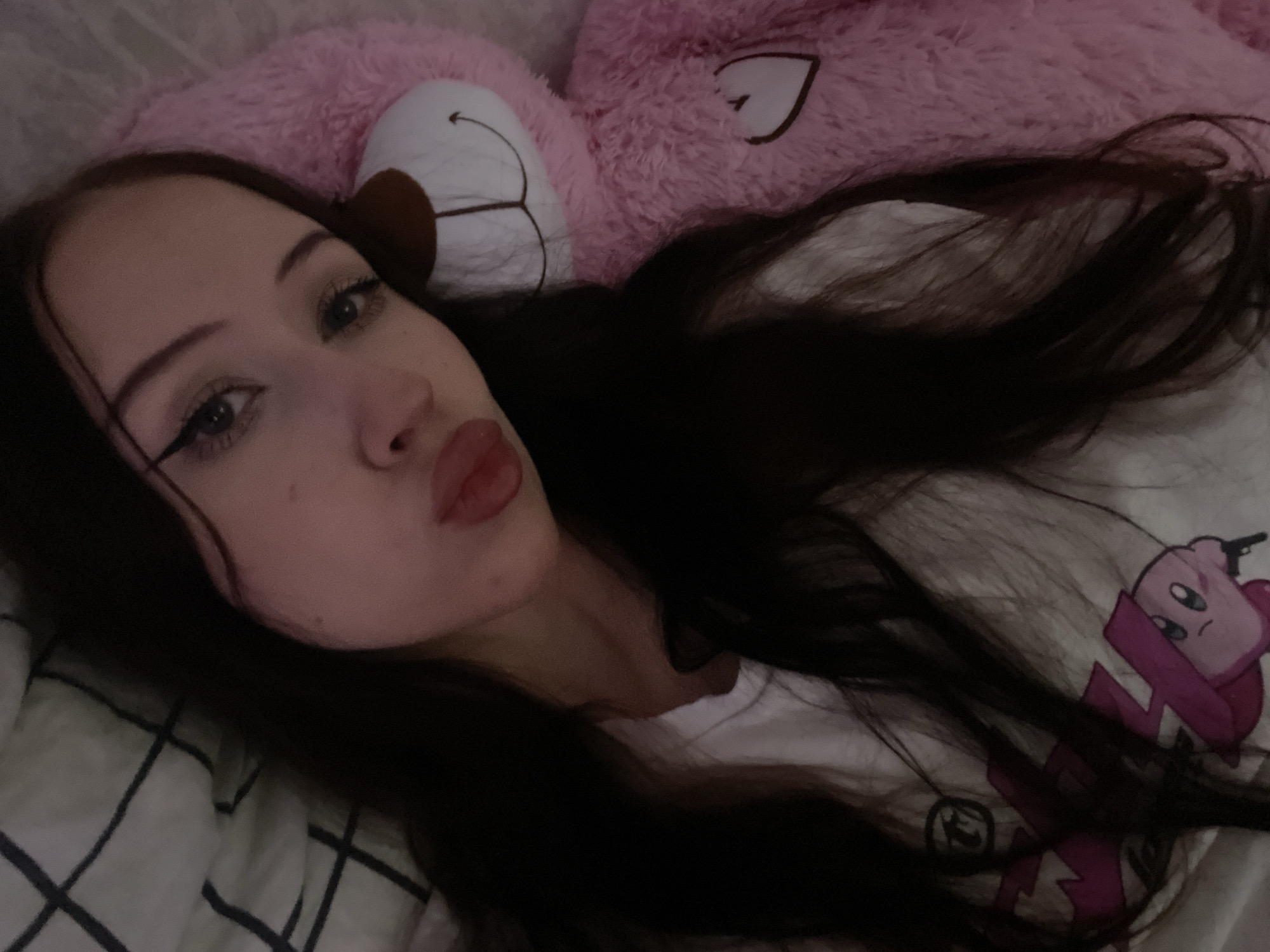 A young woman with long, dark hair is lying down next to a large, fluffy pink teddy bear. She is wearing a white t-shirt with a graphic print. Her head is tilted, and her lips are pursed in a kiss. Her eyes are open, and she is looking towards the camera. A plaid blanket is visible in the background. The scene appears to be an indoor setting, possibly a bedroom. The teddy bear has a stitched heart on its chest and a simple stitched face. The woman's makeup is noticeable, with winged eyeliner. There is no indication of the location being in Votkinsk, Russia.