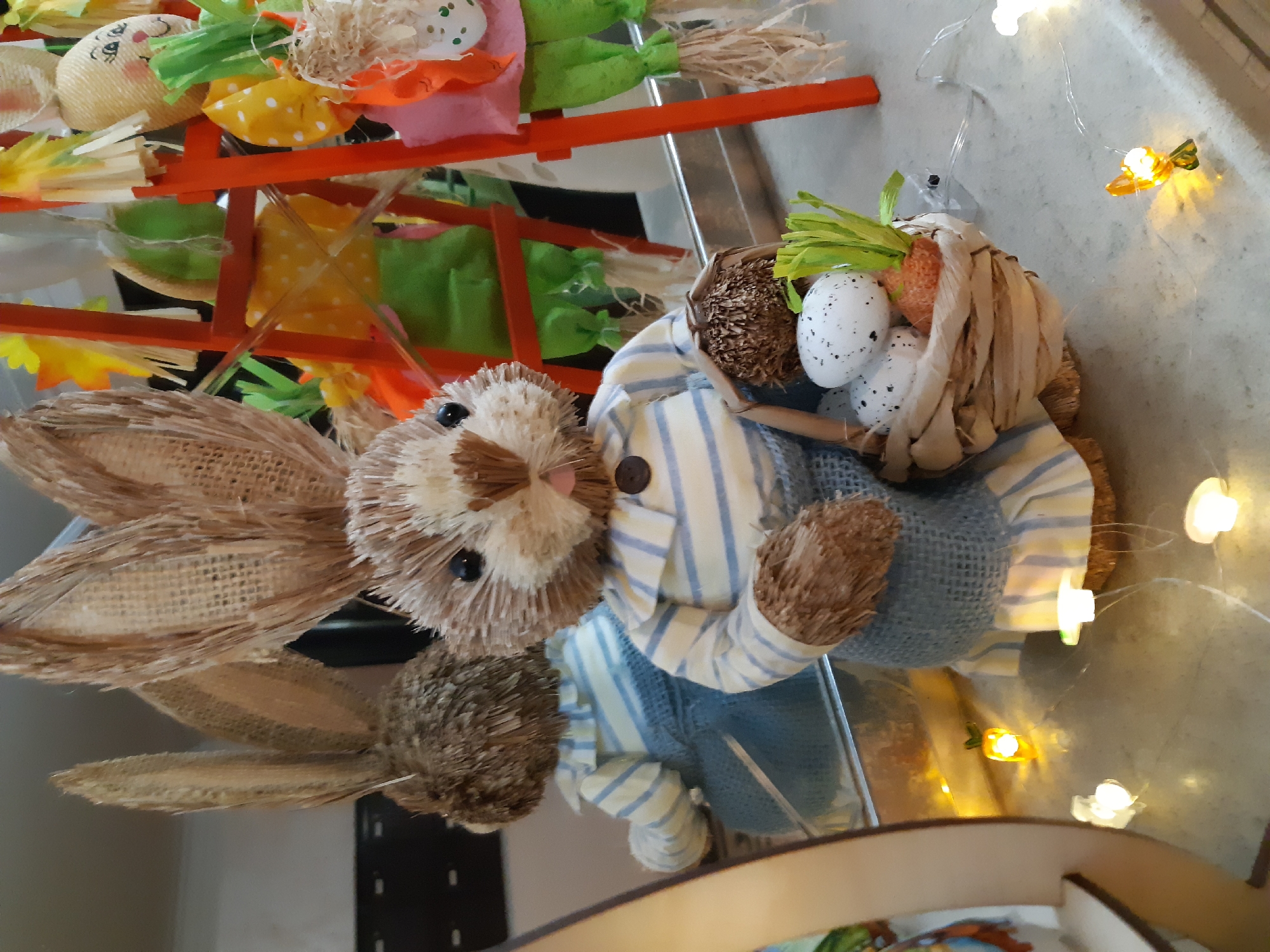 An Easter-themed scene is presented indoors. A straw-like bunny figure in blue overalls and a striped shirt is the main focus. It carries a small basket containing speckled eggs and what appears to be a carrot with green leaves. In the background, there's a tiered structure made of orange sticks holding other Easter-themed figures. Fairy lights are scattered around the scene, casting a soft glow. The reflective surface indicates that the objects are placed on a table or countertop. Location is not provided.