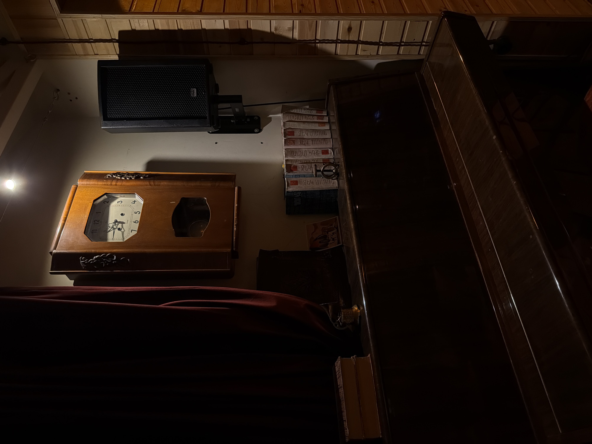 The image depicts a dimly lit interior space, likely in Yerevan, Armenia, given the presence of Armenian language packaging. A wall-mounted speaker is positioned above a wooden-framed clock. Adjacent to these is a stack of books or boxes bearing Armenian text. To the right, a dark, polished wooden cabinet or furniture piece is prominent. The lower portion of the image is obscured by a dark red, draping fabric. A faint light source is visible on the left side of the frame. The ceiling is paneled with light-colored wood.