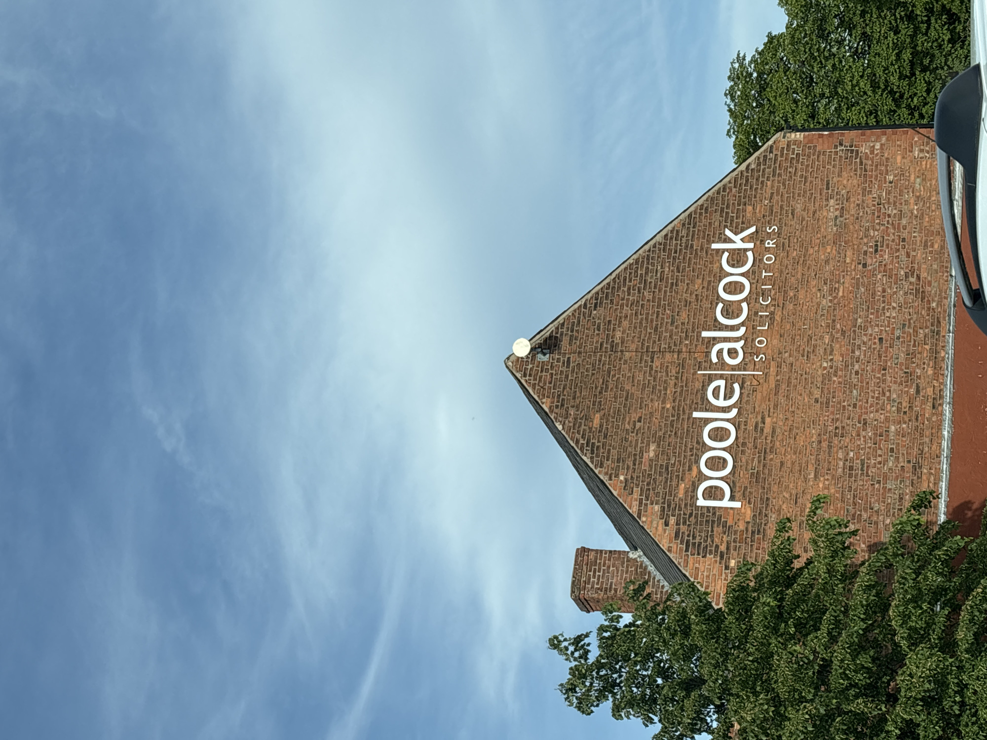 The image shows the exterior of a brick building, identified by a sign as "poole alcock solicitors," presumably located in Crewe, United Kingdom. The building has a steeply pitched roof covered in brick. A white satellite dish is mounted near the peak of the roof. Green trees partially obscure the base of the building, and a partial view of a white van or other vehicle is visible on the right side of the image. The background consists of a blue sky with wispy clouds. The photograph is taken from a low angle, looking up at the building.
