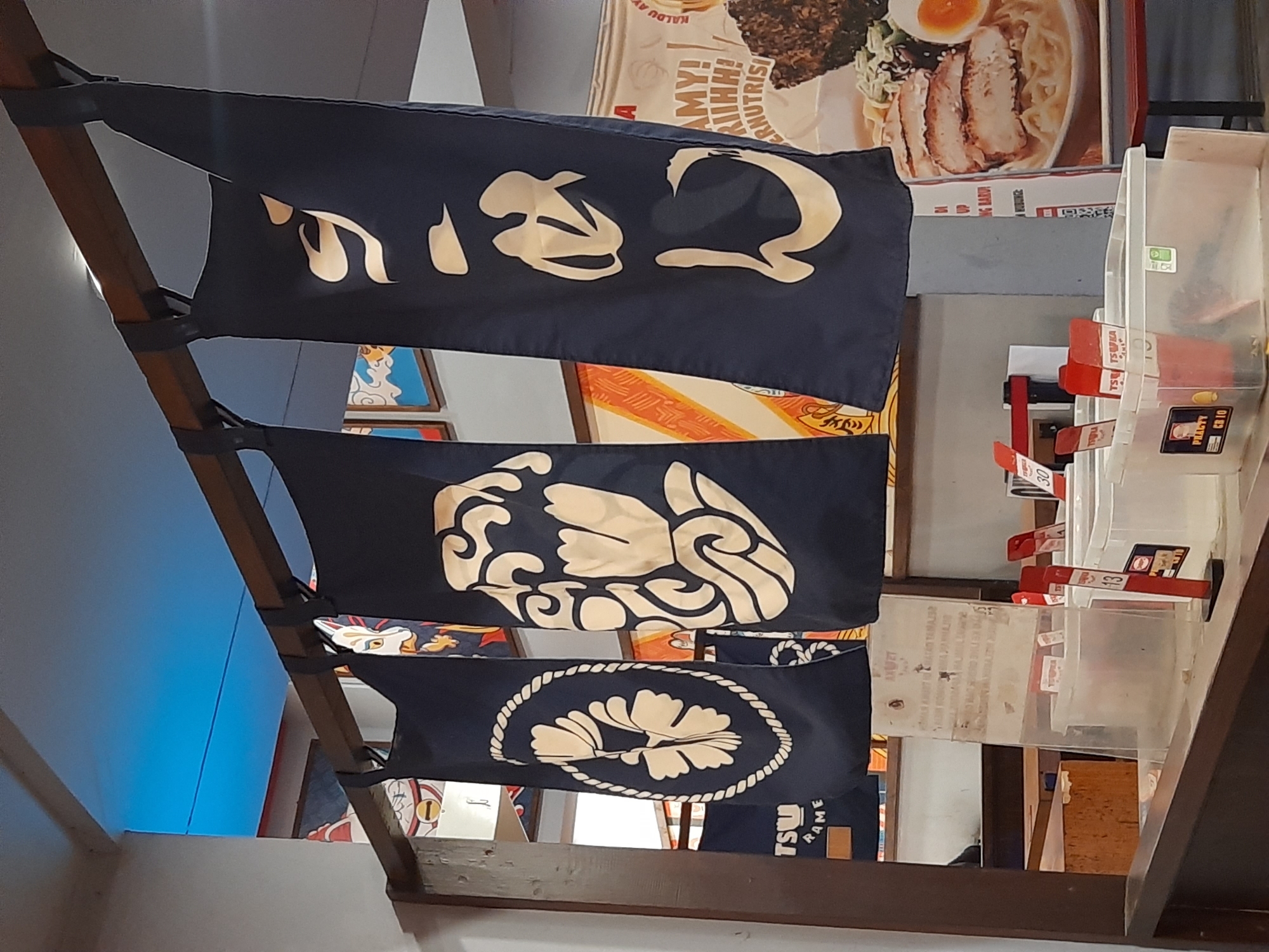 The image depicts the interior of a ramen restaurant, likely located in Bandung City, Indonesia, as suggested by the presence of Japanese cultural elements and a general ambiance found in such establishments in the city. The foreground features a counter area with various plastic containers holding utensils and condiments, some with labels such as "PHALTY CB 10" and "TSUKU RAMEN". Red holders with numerical tags like "13" and "30" are also visible. Behind the counter, four dark blue fabric banners with large, light-colored Japanese calligraphy or symbols hang from a wooden beam. The banners appear to be traditional noren curtains, common in Japanese eateries. One banner on the far left displays a circular emblem with a floral design, while the second from the left shows a stylized wave-like pattern. The third banner features a face, possibly a Kitsune mask, and the fourth displays more Japanese text. In the background, tables and chairs are arranged, and a large promotional poster showcasing a bowl of ramen with chicken and an egg is visible on the right. The lighting suggests an indoor setting, with some blue lighting visible near the ceiling on the left. The overall scene conveys a casual dining environment focused on Japanese cuisine.