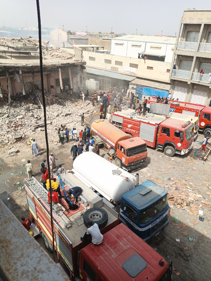 The image depicts an emergency scene in Fagge C, Nigeria, showing the aftermath of a structural collapse or significant incident. The foreground features multiple emergency vehicles parked on a debris-strewn ground. Three red fire trucks are prominent; one in the immediate foreground has a person in a white shirt sitting on its roof, while others are seen on its flatbed, some in red and yellow shirts, operating equipment or hoses. Another red fire truck is positioned further back, with hoses deployed. An orange tanker truck and a white tanker truck are also present, with individuals near them.
To the left, a multi-story building is severely damaged, with its upper floors having collapsed into a large pile of rubble at ground level, exposing internal structures and debris. To the right of the scene, an adjacent multi-story building appears structurally intact, with several individuals visible on its balconies.
A large crowd of civilians and emergency personnel is gathered in the central area, amidst the debris and around the emergency vehicles. Smoke or dust is visible in the background, rising from the collapsed structure. The ground is covered with rubble, bricks, and discarded items. The overall scene indicates ongoing emergency response efforts in an urban environment under daylight conditions.