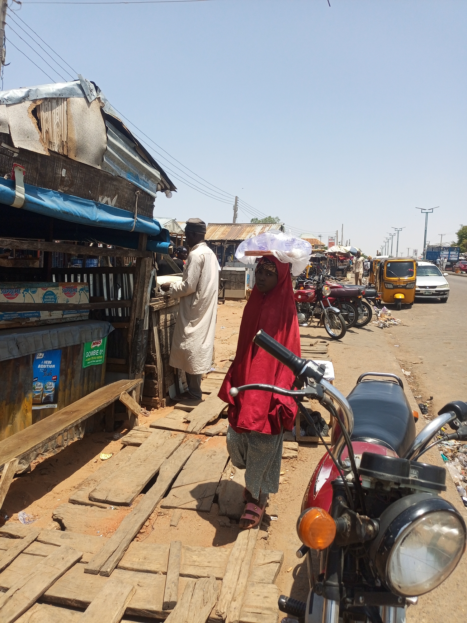 This image captures a busy street scene in Gombe, Nigeria, under a bright, clear sky, suggesting daytime, possibly midday or early afternoon. The ground is a mix of dirt and uneven wooden planks, typical of a market or roadside stall area.
In the center foreground, a woman wearing a red hijab and grey patterned trousers stands, balancing a white package on her head. She appears to be looking towards the left side of the frame. To her immediate left, an older man in a light-colored long garment and a cap is attending a wooden stall. The stall is constructed from raw wood and corrugated metal sheets, with a blue tarp serving as an awning.
Visible items on the stall include multiple boxes labeled "SUPREME NOODLES, TASTY CHICKEN INSTANT NOODLES," with additional text "40 Packs x 120g." Below these, a poster for "Peak" milk advertises "A NEW ADDITION TO Peak." Another poster advertises "MILO" and mentions "GOMBE 01 $100." A simple wooden bench is attached to the stall's front.
To the right of the woman, the front section of a red motorcycle, including its headlight and handlebars, is prominent in the foreground. In the mid-ground, behind the woman, more motorcycles are parked. A yellow commercial tricycle (Keke Napep) and a white car are visible on a paved road, indicating active traffic. The background shows more market structures, people, utility poles, and modern streetlights, suggesting a bustling urban or semi-urban commercial area. The overall scene depicts daily commerce and street life in the region.