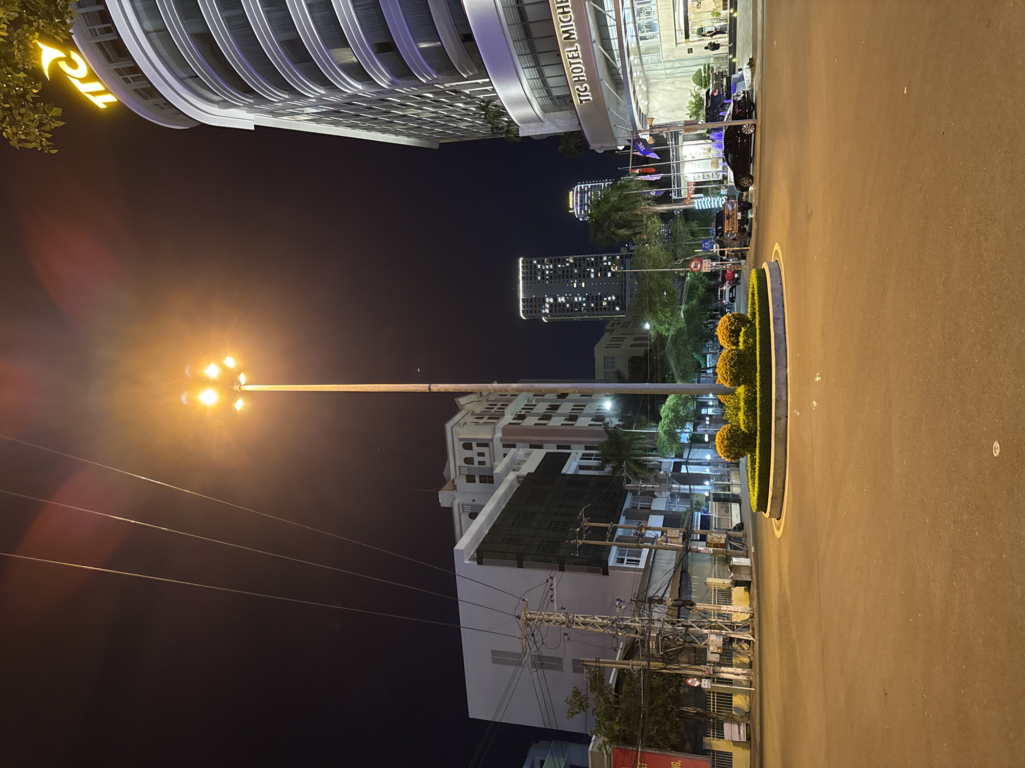 The image depicts a nighttime street scene in Nha Trang Ward, Vietnam. The primary subject is a modern building complex, with a prominent curved structure featuring a bright yellow "ITC" logo on its facade. Next to it, another building has "TTC HOTEL MICHEL" written in white text. The scene is illuminated by streetlights, including a tall lamppost with multiple bright yellow lights that cast a warm glow on the surrounding area.
In the background, several tall buildings with many illuminated windows are visible, suggesting an urban environment. Palm trees and other greenery are scattered throughout the scene, adding to the tropical feel often associated with Vietnam. The road surface is asphalt, with a yellow line marking the edge of a landscaped median. This median features neatly trimmed bushes and small, round, orange-flowered plants.
A few cars are parked or driving on the road. One dark-colored SUV is parked near the hotel entrance, and a darker vehicle is partially visible further down the street. There are also electrical wires and poles crisscrossing the frame, indicating utility infrastructure.
The time of day is clearly night, with a dark sky and artificial lighting providing illumination. The weather appears clear, as there are no visible signs of rain or fog. The overall impression is one of a well-lit, modern urban setting at night, likely in a tourist or commercial district. No specific event is taking place; it's a general view of the street.