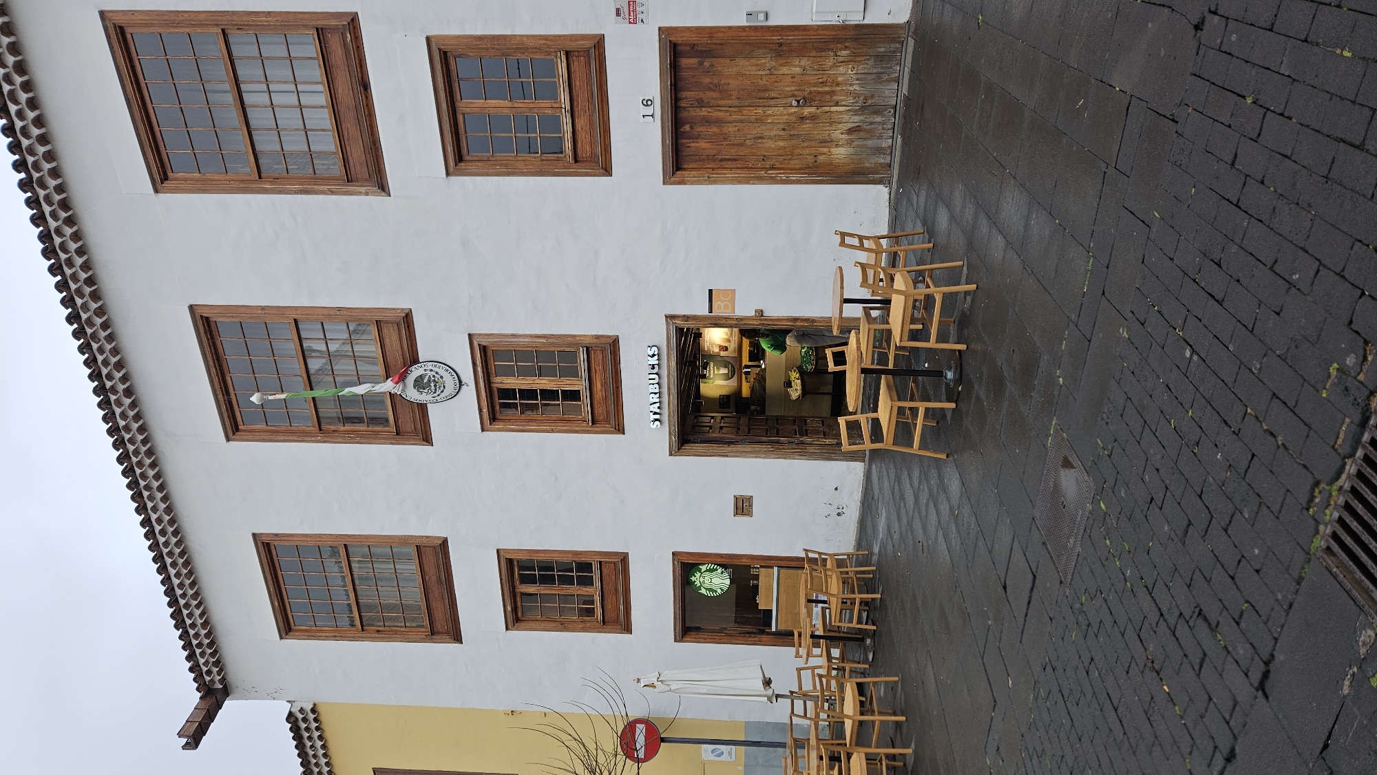An image captured from a street level depicts the facade of a two-story building with a white stucco exterior and traditional wooden windows and doors. The building, numbered "16" to the right of a large double wooden door on the upper floor, occupies the left side of the frame. Below this door, there's another entrance framed by dark wood, leading into what is clearly identifiable as a Starbucks coffee shop, with "STARBUCKS" written above its awning and the green Starbucks siren logo visible near another lower entrance.
The building features several rectangular wooden windows with multiple panes. On the upper floor, two large windows are visible, and on the lower floor, there are four smaller windows. A distinctive banner, green, white, and red, hangs from one of the lower-level windows, featuring a circular emblem with what appears to be a coat of arms or official seal and some illegible text. An alarm box is visible on the white wall near the upper right window.
In front of the building, a dark, cobblestone or brick-paved street stretches out, appearing wet, suggesting recent rain. Patches of green moss or growth are visible in the cracks of the paving. Scattered across the wet pavement are numerous light-colored wooden chairs and small round tables, arranged in groups, typical of an outdoor cafe seating area. Most chairs are empty, and some are stacked or pushed against tables. A drain grate is visible on the far right of the street, and a square metal cover is embedded in the pavement closer to the building.
The lower left corner of the image shows the top of an adjacent yellow building with a similar tiled roofline, and a red circular road sign (possibly a "No Entry" or "Stop" sign) is partially visible on the street level next to an outdoor umbrella stand. The scene appears to be set during daytime, likely overcast given the soft, even lighting and the wet ground. The architecture and street scene are consistent with the historical context of San Cristóbal de La Laguna, Spain.