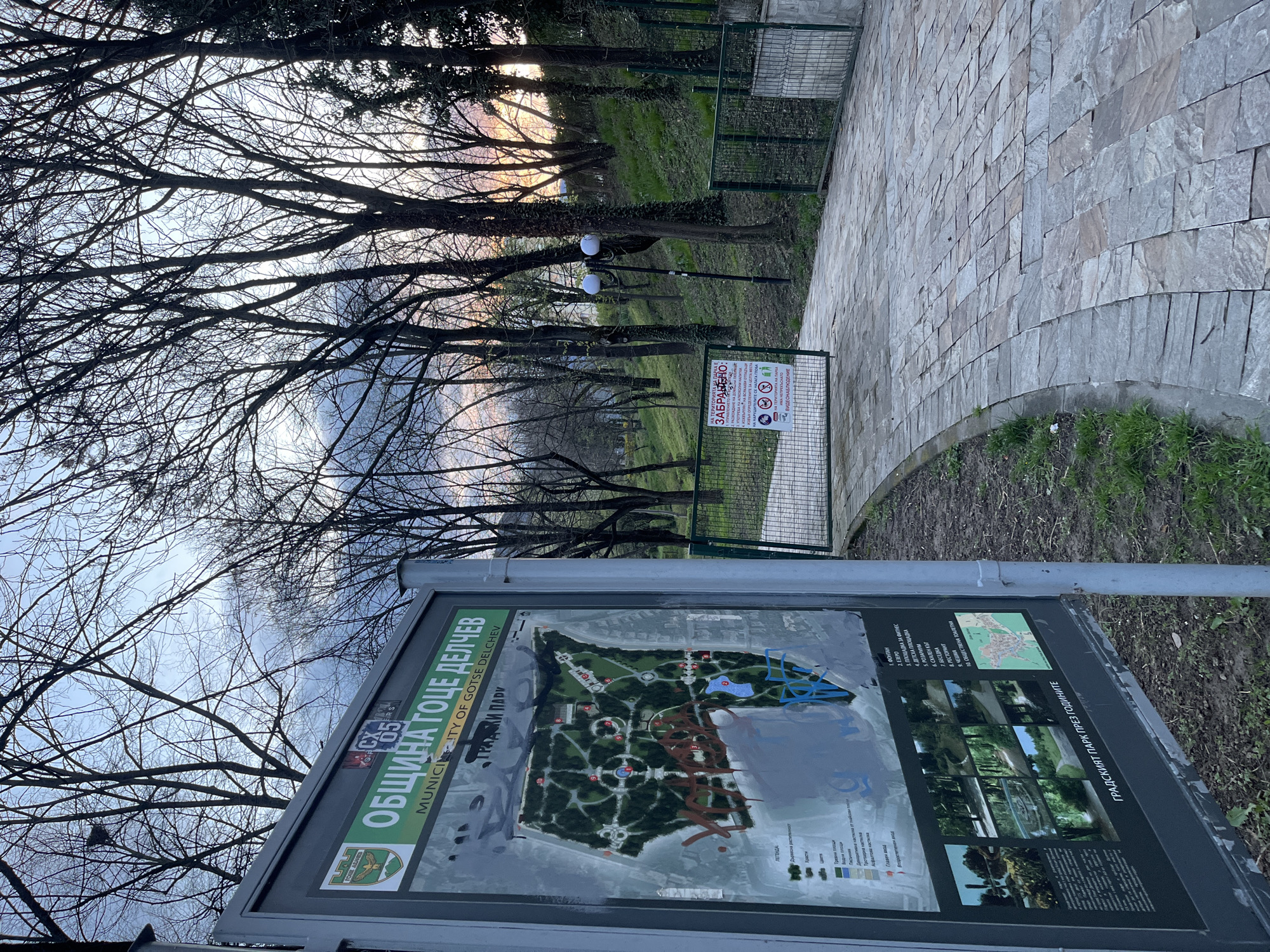 The image shows a park scene in Goce Delcev, Bulgaria, with a map of the park in the foreground. The map is partially obscured by graffiti. Behind the map, a pathway leads into the park, with trees lining either side. A sign with prohibitions is visible along the path. The trees are mostly bare, suggesting it is either late autumn or early spring. The sky is overcast with hints of sunset.