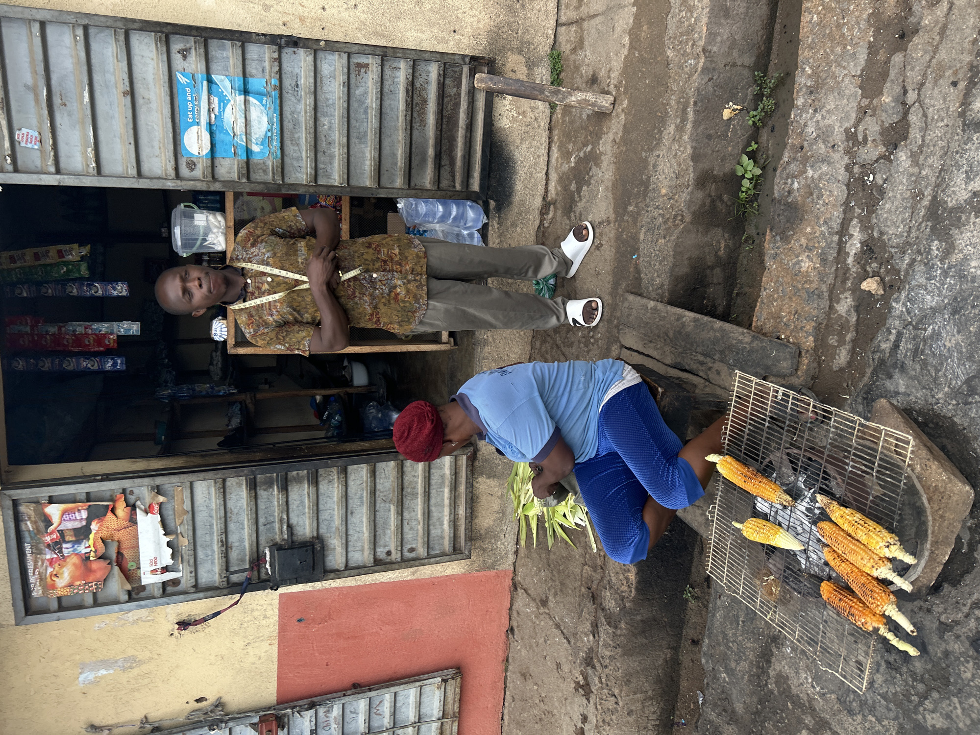 The image shows a street scene in Jos, Nigeria. A man stands in front of a corrugated metal storefront with a partially open door. The storefront displays various items. He is wearing a patterned shirt and khaki pants, with white sandals. A tape measure is draped around his neck. Outside the store, a woman is kneeling and tending to corn on a grill, set on a charcoal fire. She is wearing a blue shirt and blue leggings. The ground is paved with concrete.